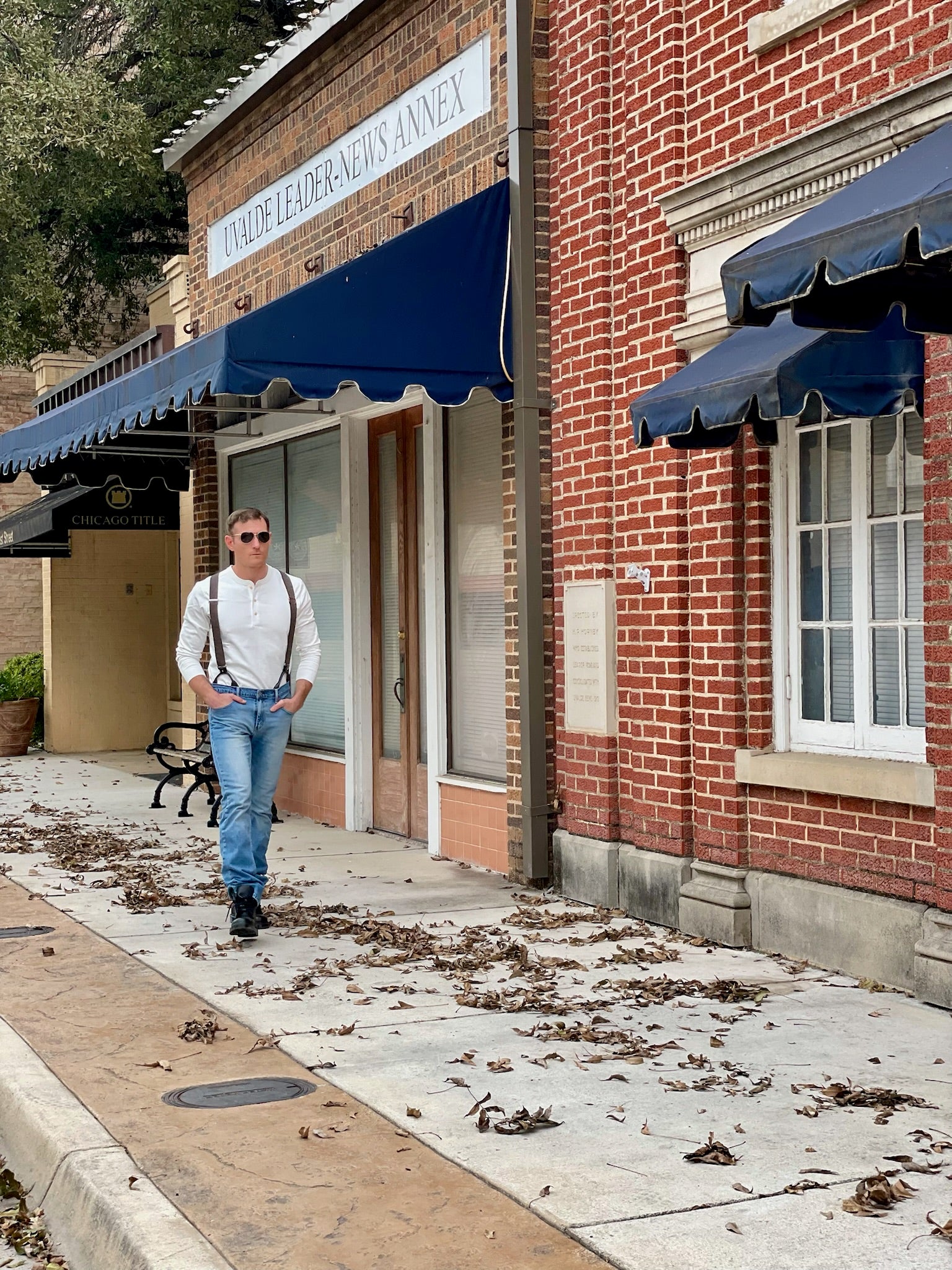 A man walks through a historic downtown wearing Espresso Linen Suspenders with black leather button-on attachments, paired with a vintage white Henley and classic denim. Handmade in Texas by Stratton Suspender Co., perfect for weddings, formalwear, and heritage-inspired everyday menswear.
