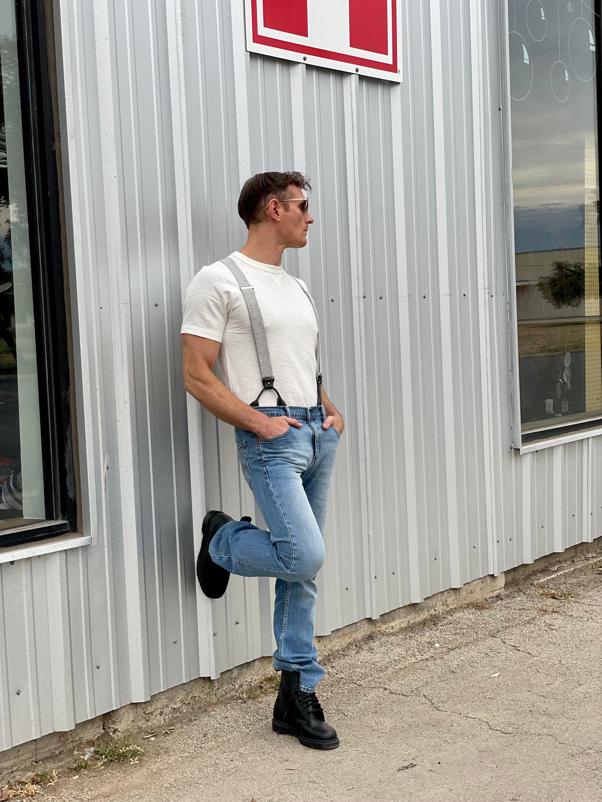 A stylish man wearing gray linen button-on suspenders paired with a classic white t-shirt, light-wash denim jeans, and black leather boots, leaning casually against a corrugated metal wall. These handmade suspenders feature full-grain leather button tabs and premium metal hardware, offering a timeless look perfect for weddings, groomsmen, and refined vintage menswear.
