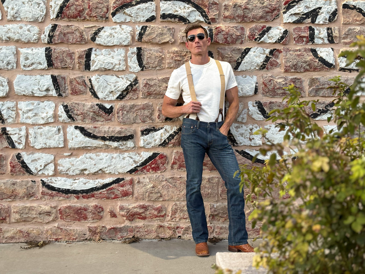 A man wearing a white t-shirt, blue jeans, beige suspenders, and brown leather shoes stands against a rustic brick wall with a painted mural. A green leafy plant is visible in the foreground on the right.