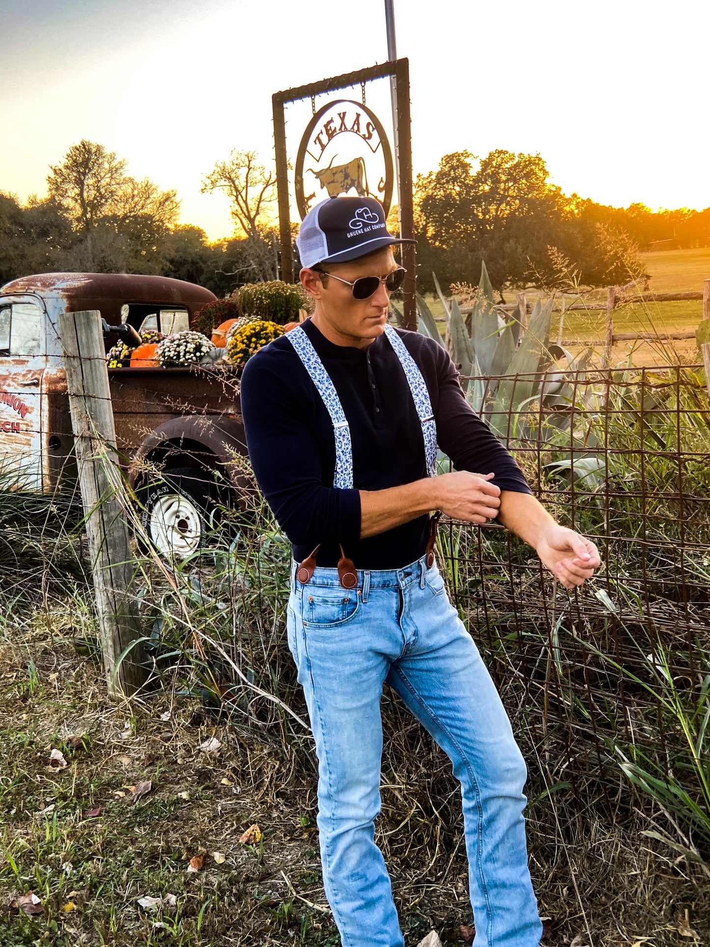 A groom styling Sapphire Blue Floral wedding suspenders with a navy blue Henley shirt and light-wash jeans at a rustic Texas wedding. These button-on suspenders feature a vintage-inspired blue floral pattern on linen straps, full-grain leather button-on attachments, and polished brass hardware. Designed for grooms, groomsmen, and rustic wedding themes, offering a refined, tailored look superior to clip-ons. Perfect for country weddings, vintage menswear, and heritage-inspired formalwear.