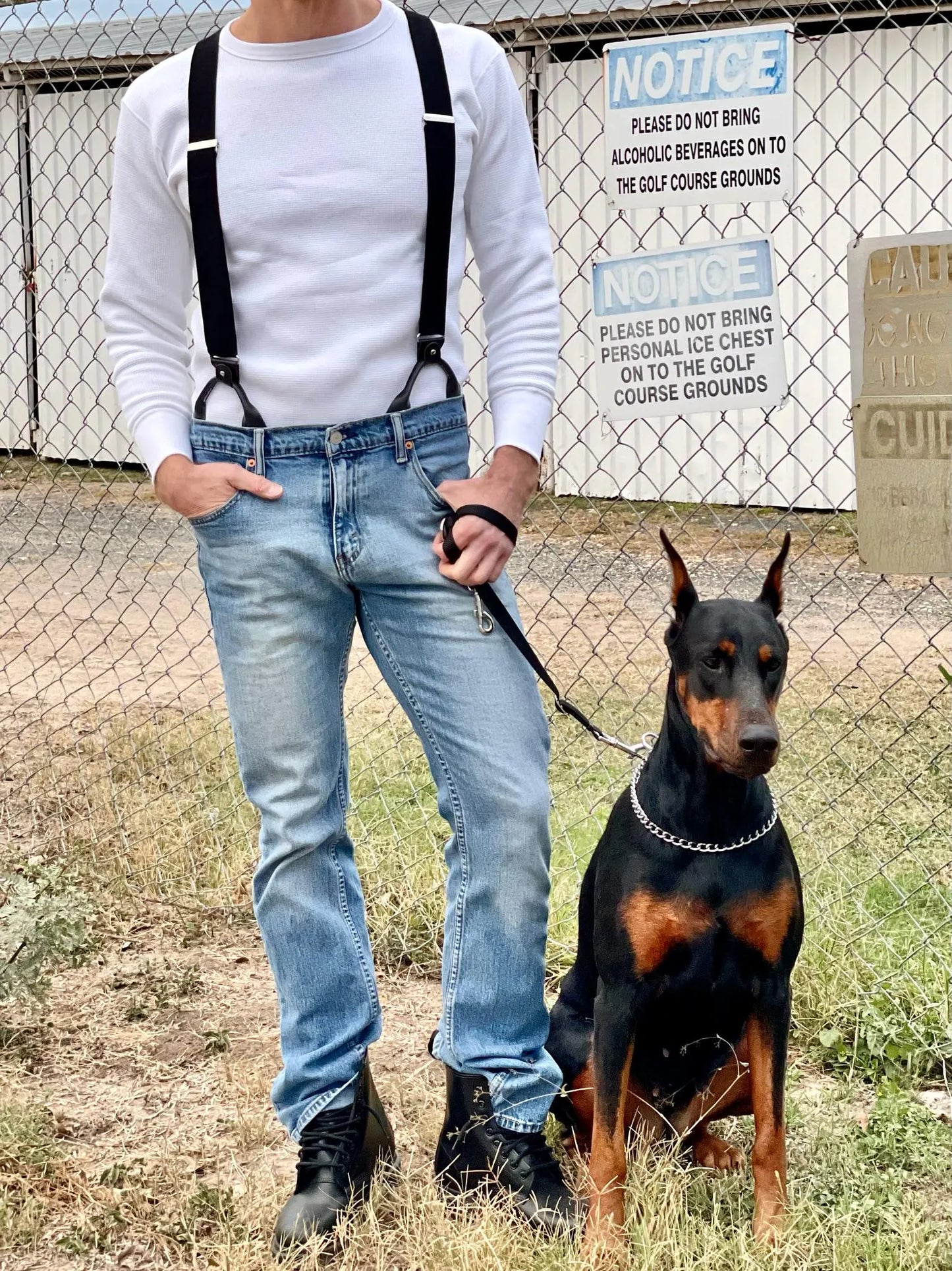 Man wearing black linen button-on suspenders by Stratton Suspender Co. paired with a white waffle-knit shirt and Levi’s jeans, standing with a Doberman against a chain-link fence—rugged handmade accessories crafted in Austin, Texas.