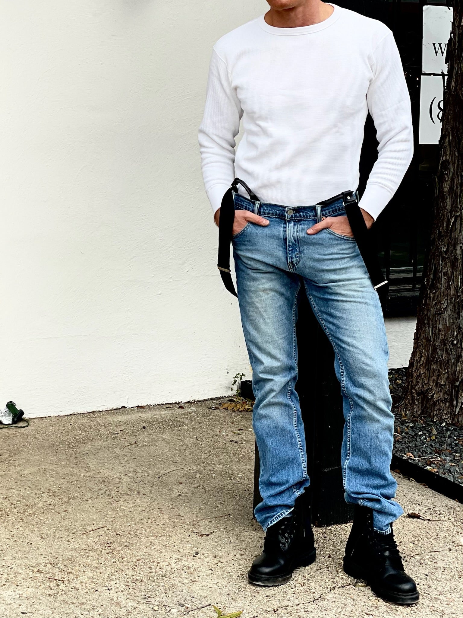 Men’s black suspenders styled hanging down with faded blue jeans, combat boots, and a white thermal shirt for a rugged everyday look.