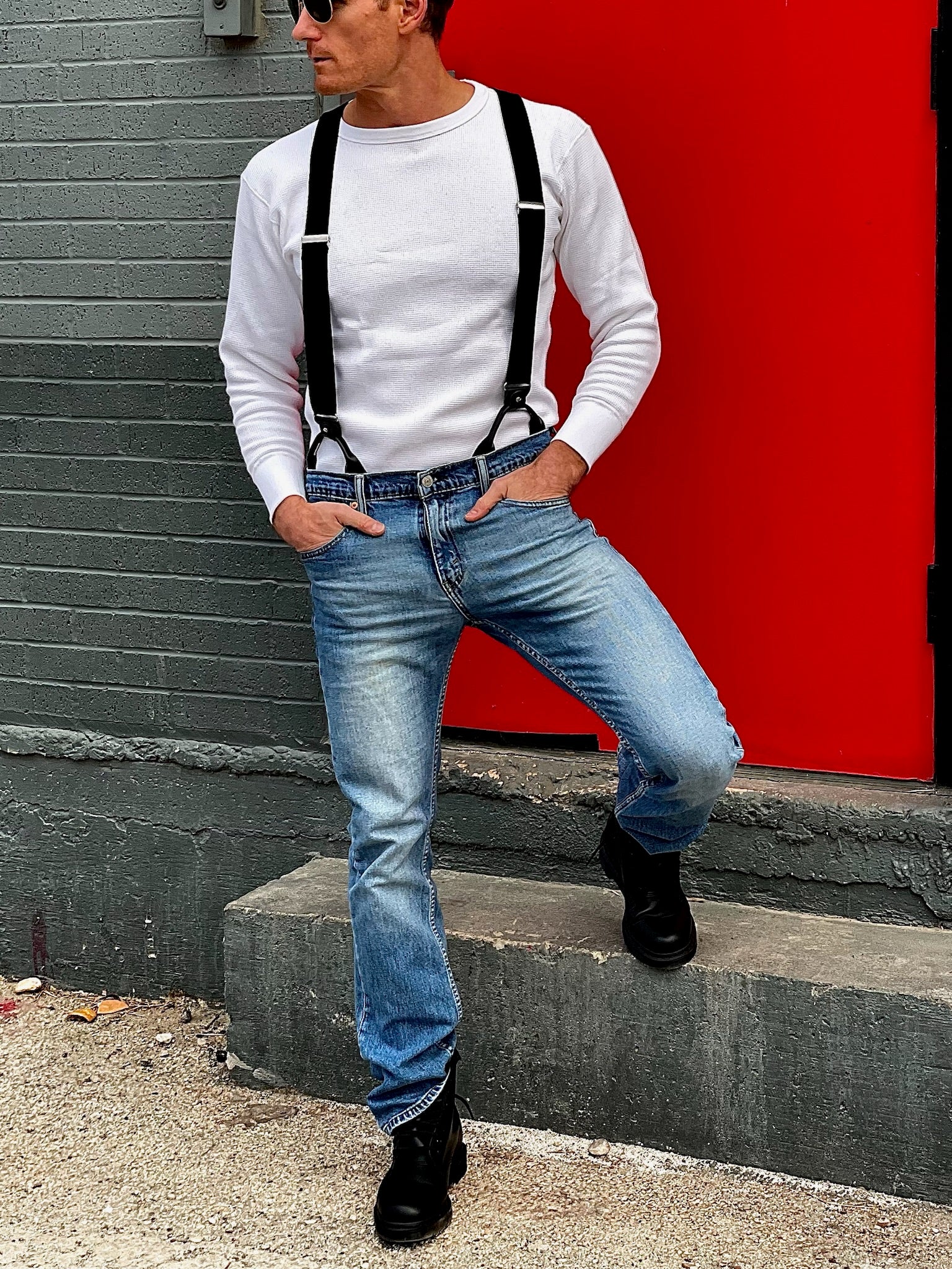 Model wearing black linen button-on suspenders with silver hardware, styled with light wash Levi’s jeans and a white waffle knit shirt, standing against a bold red and gray industrial backdrop—handmade by Stratton Suspender Co. in Austin, Texas.