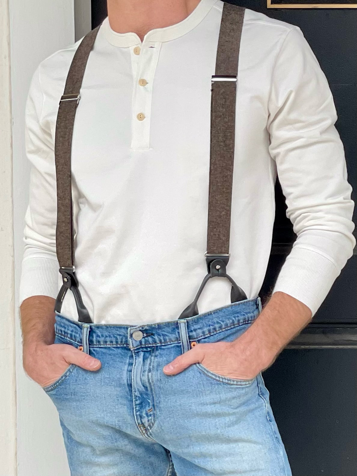 Close-up of man wearing brown rustic suspenders with black leather button-on loops over a cream henley shirt and light-wash jeans — handcrafted men’s suspenders for casual or rugged everyday style.
