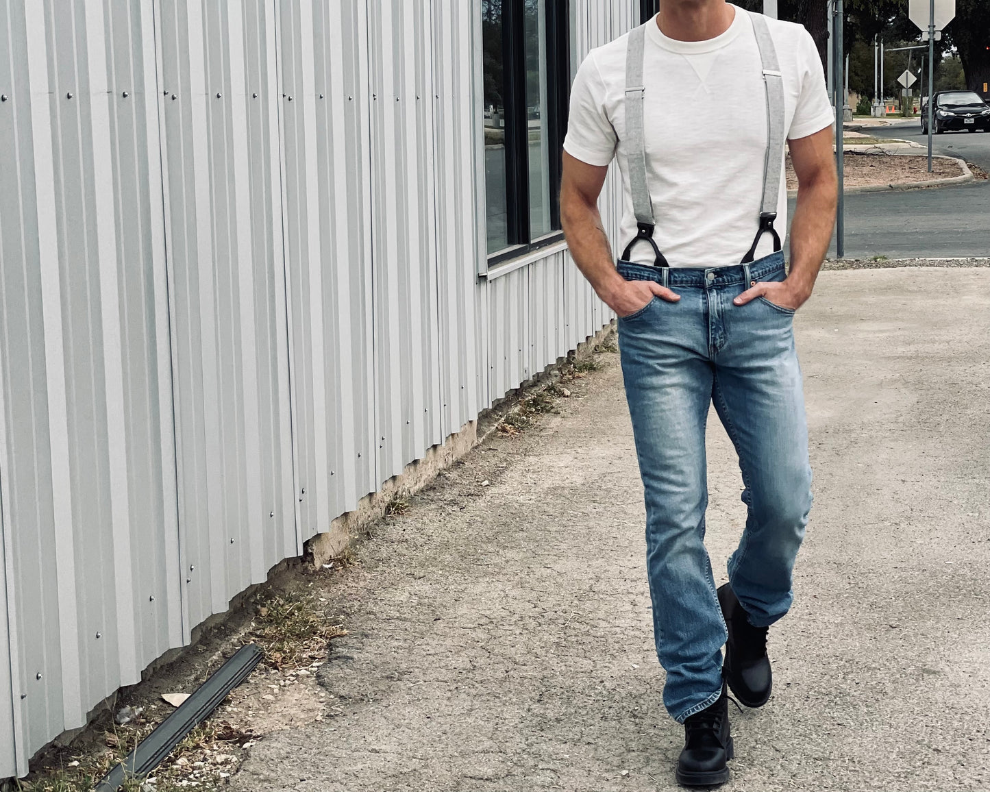Man walking confidently in charcoal and beige woven linen suspenders with leather button tabs, styled with a white tee, light-wash jeans, and black boots, in front of corrugated metal siding for a rugged-modern Texas look.