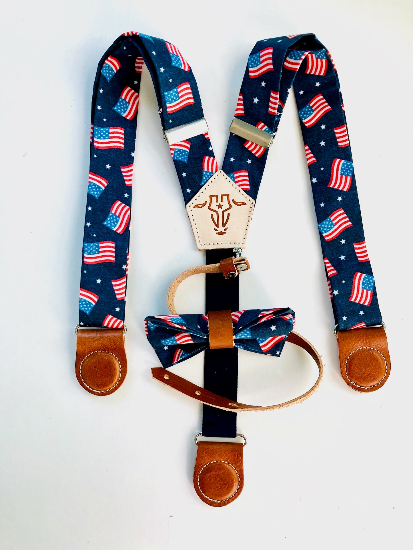 Kids’ patriotic suspenders featuring a USA flag print, tan leather magnetic clasps, and a coordinating bow tie for Independence Day events.
