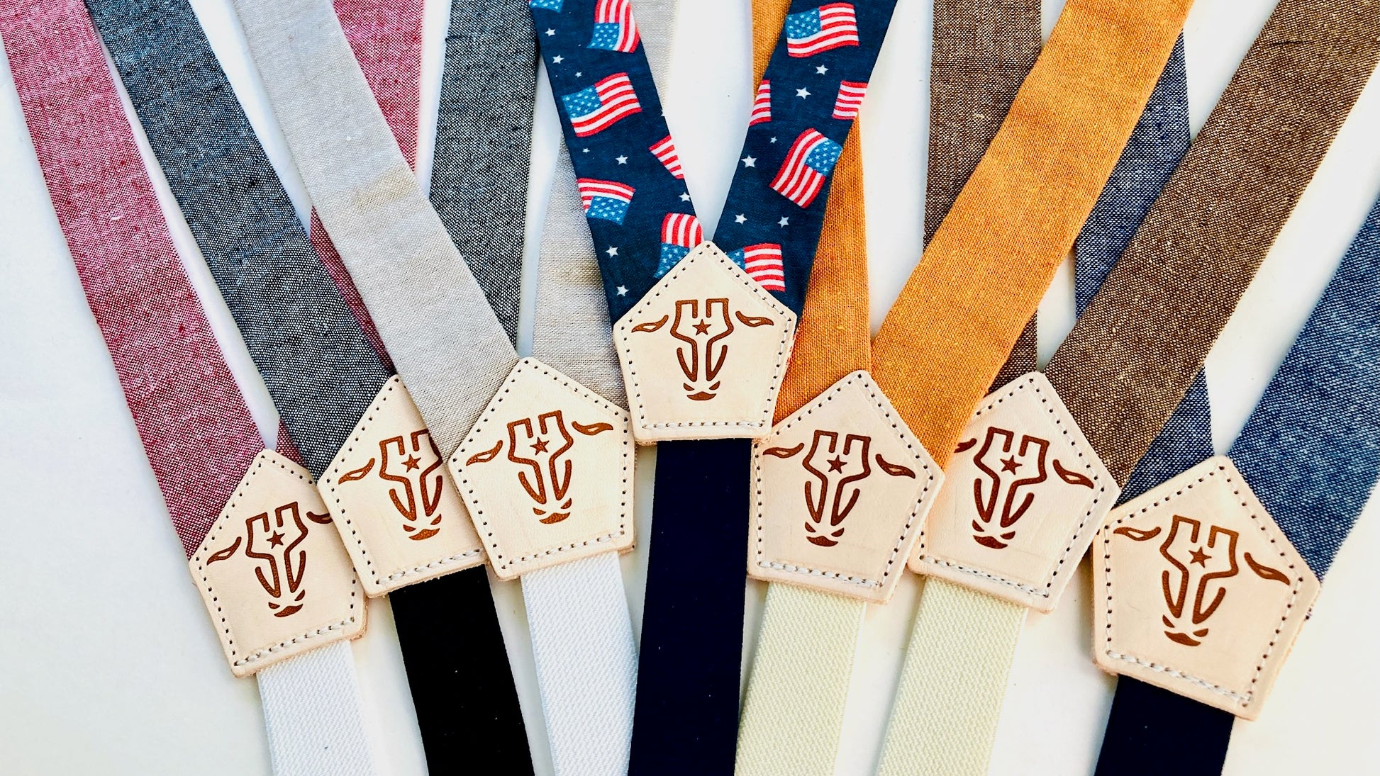Children’s suspender collection in red, gray, beige, blue, brown, orange, and patriotic flag print, displayed with stitched leather back patches.