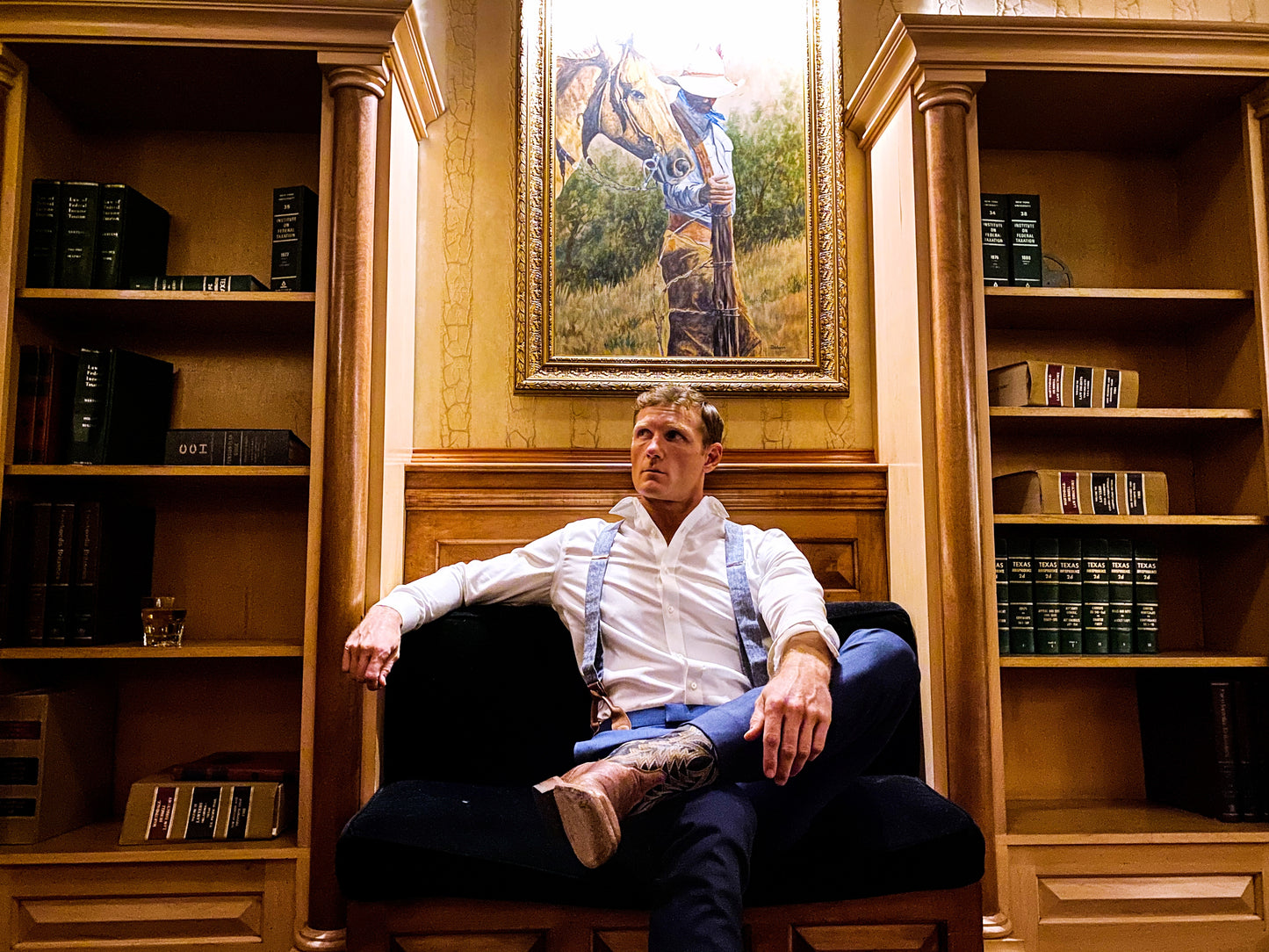 Groom or groomsman seated in the Lounge Bar at the Driskill Hotel in Austin, Texas, wearing Stratton Suspender Co. light blue linen suspenders with leather button ends and western boots for a formal rustic wedding look.
