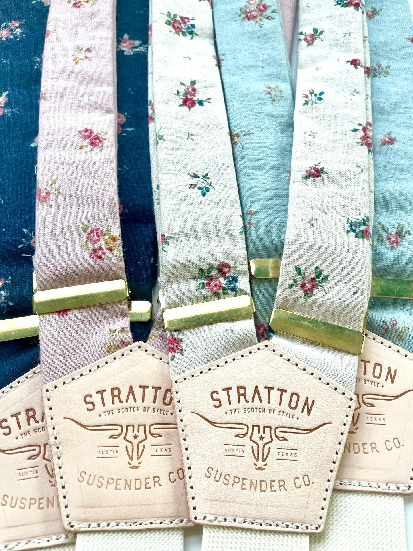 Lineup of Stratton Suspender Co. handmade floral linen suspenders in blush, navy, ivory, and powder blue with gold adjusters and tan leather back patches, perfect for weddings and groomsmen.
