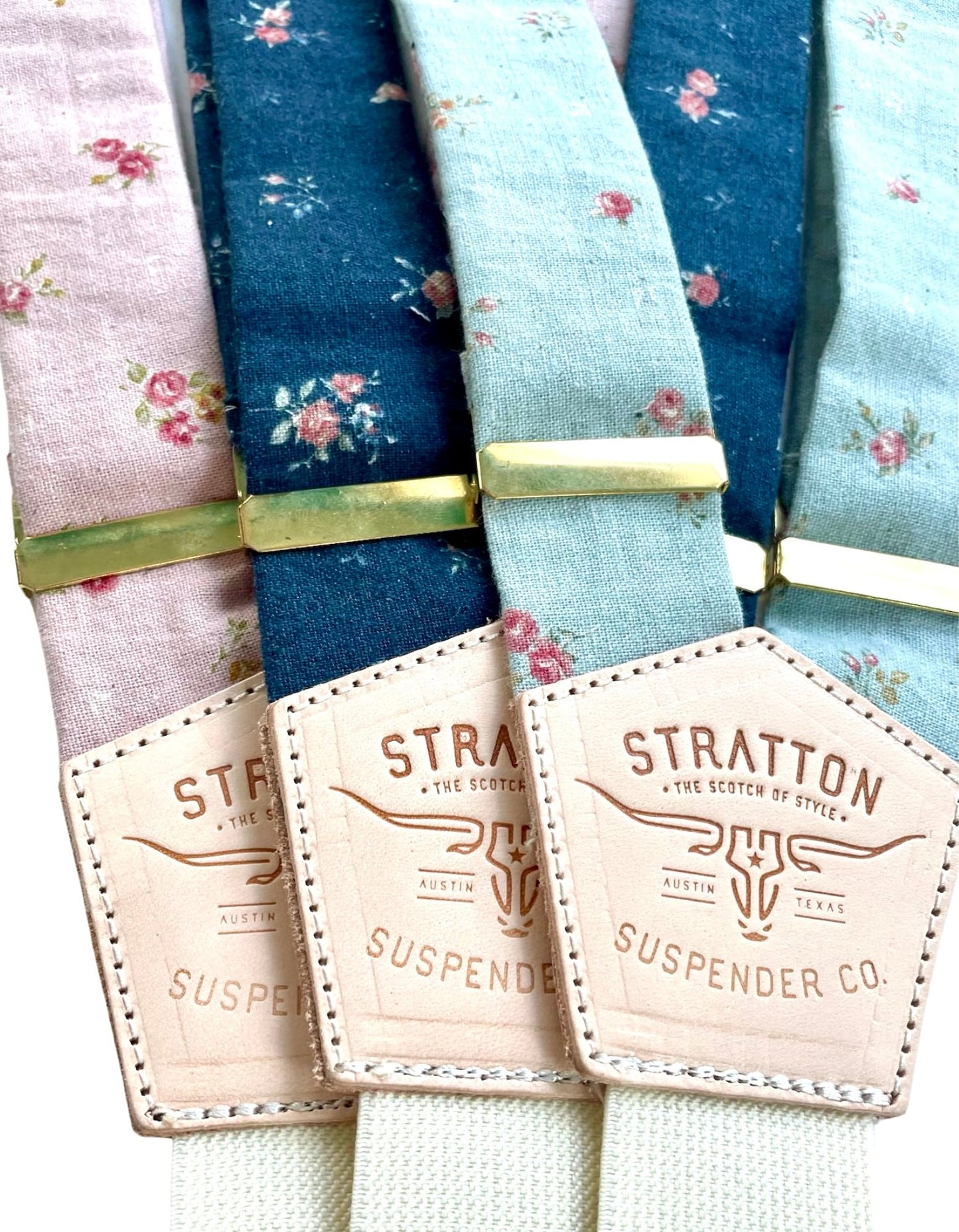 Close-up of three pastel floral linen suspenders from Stratton Suspender Co., featuring light pink, navy, and powder blue patterns with natural leather back patches—perfect for rustic or garden weddings.