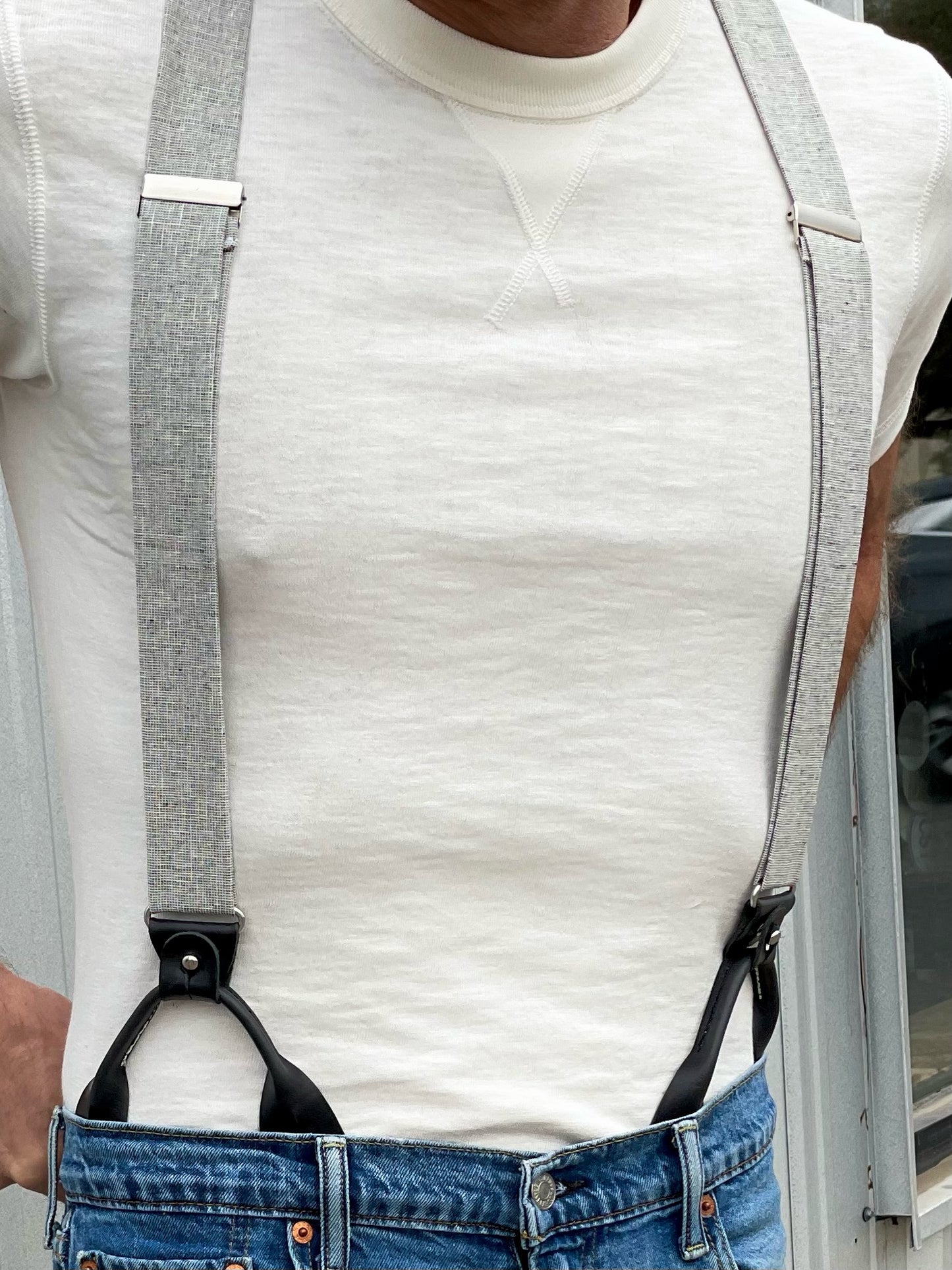 Close-up of gray linen button-on suspenders with black leather tabs by Stratton Suspender Co., shown with a cream tee and denim jeans. Handmade in Texas for rustic weddings and American menswear style.