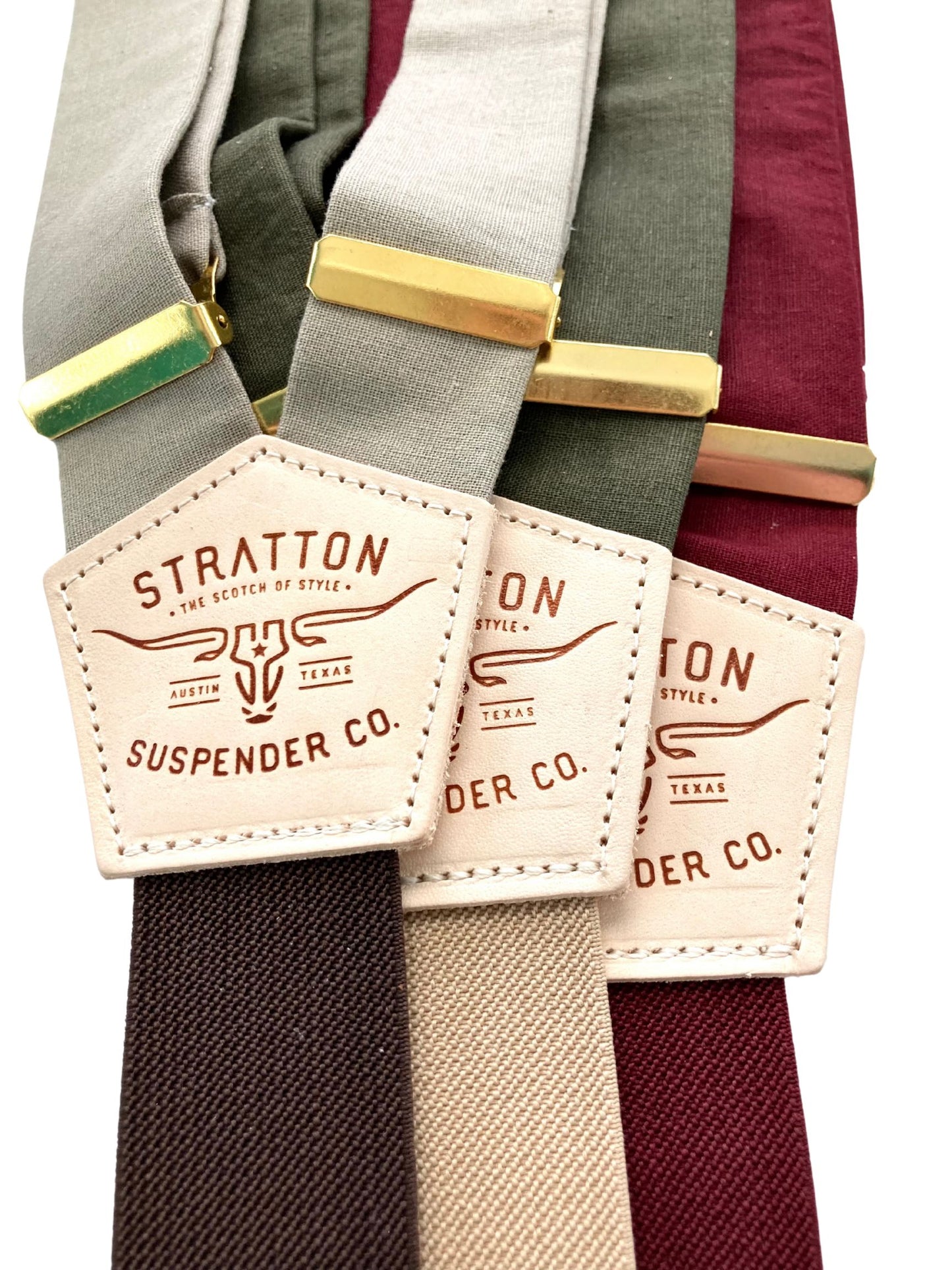 Close-up of Stratton brand suspenders in burgundy, olive green, and mushroom taupe with gold hardware and leather logo patch.