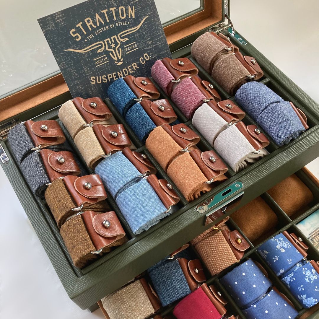 Display case of handmade linen button-on suspenders with leather attachments by Stratton Suspender Co., featuring assorted colors and textures for weddings and heritage menswear.