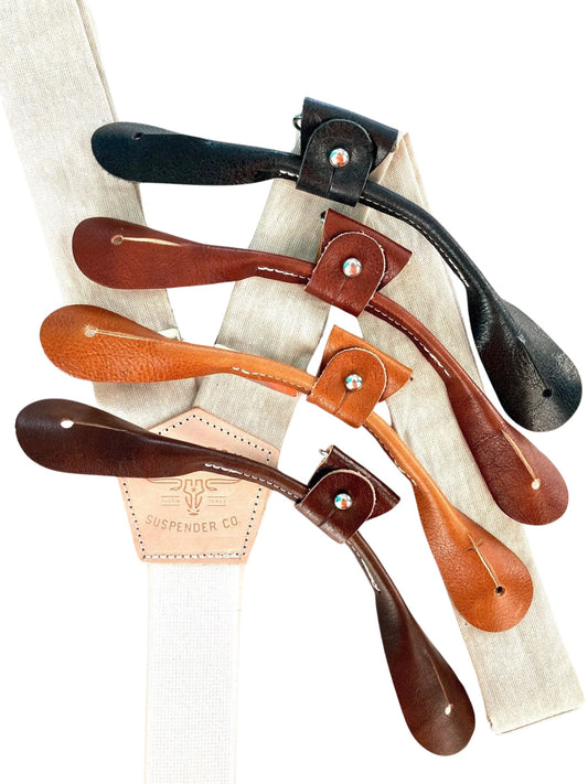 Ivory beige linen suspenders with black, cognac, chocolate, and tan leather button-on attachments arranged in a fan layout. Handmade in Texas by Stratton Suspender Co., ideal for rustic weddings, groomsmen, and vintage-inspired formalwear.