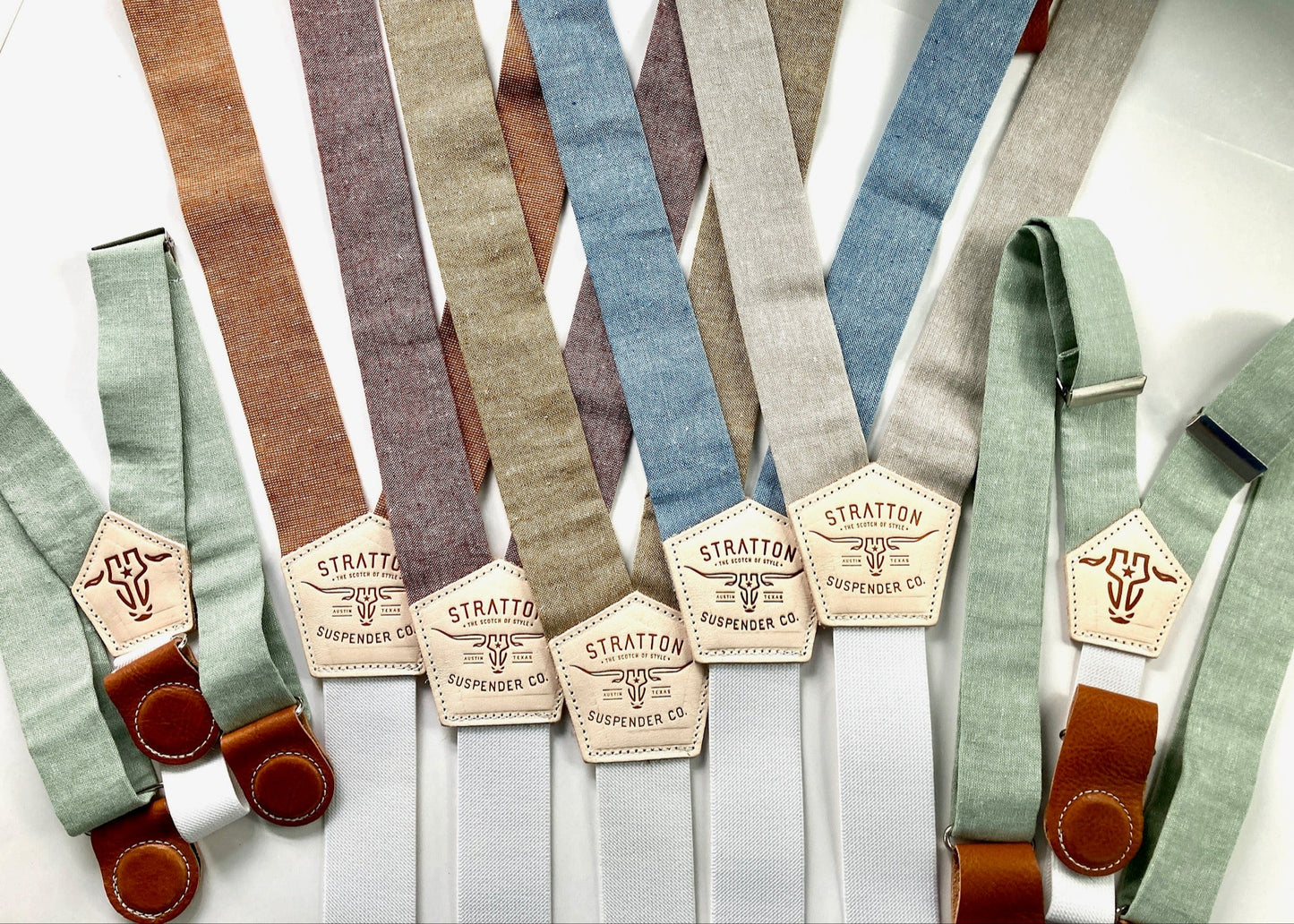 Flat lay of Stratton Suspender Co. men’s linen suspenders in sage green, burnt sienna, dusty rose, taupe, blue, and cream, with button and clip attachments, ideal for rustic or modern weddings.