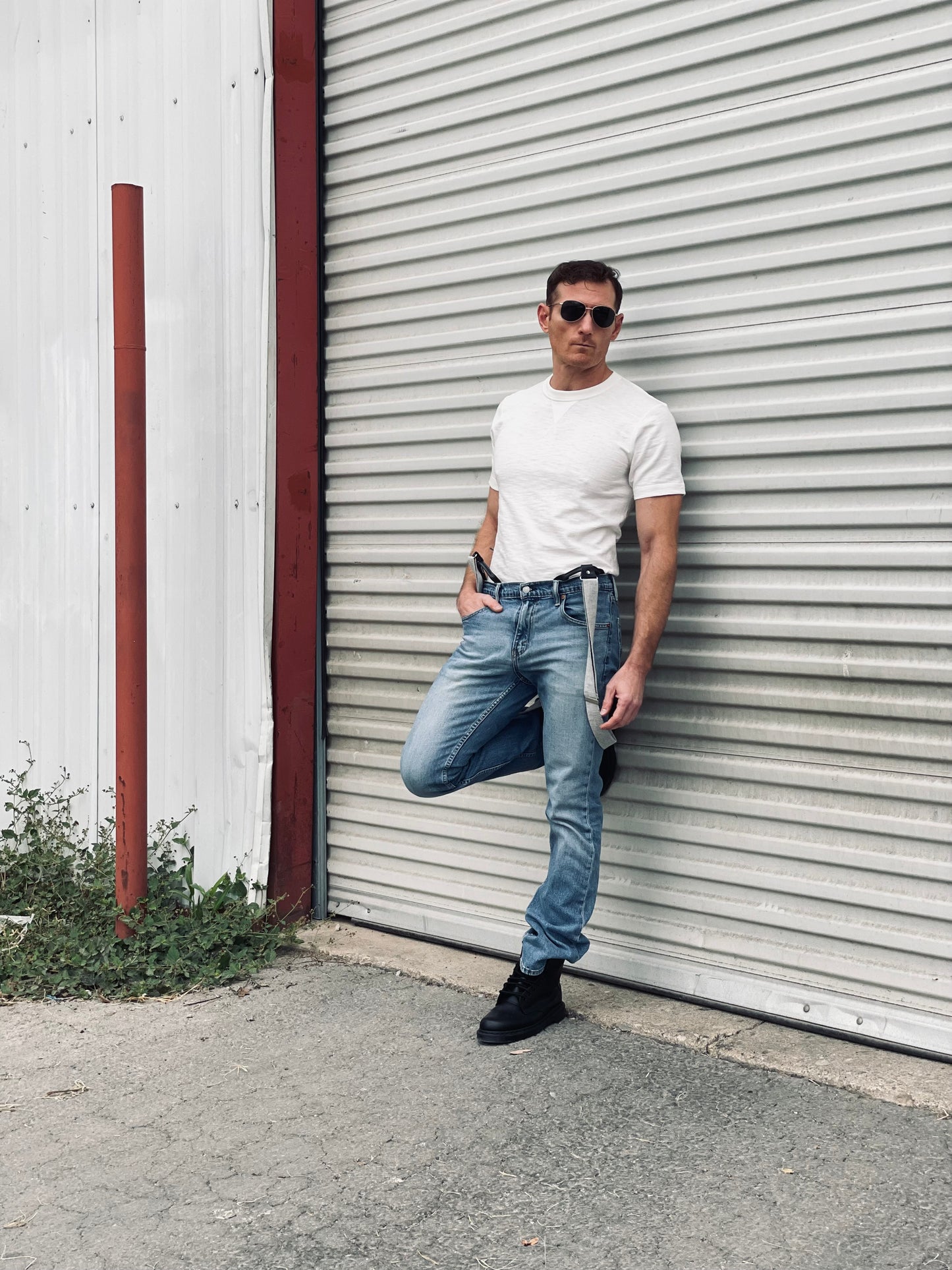 Man wearing light gray canvas suspenders with leather loops, blue jeans, and a white tee, standing outdoors against a corrugated metal wall — rugged men’s casual suspenders for everyday or workwear style.