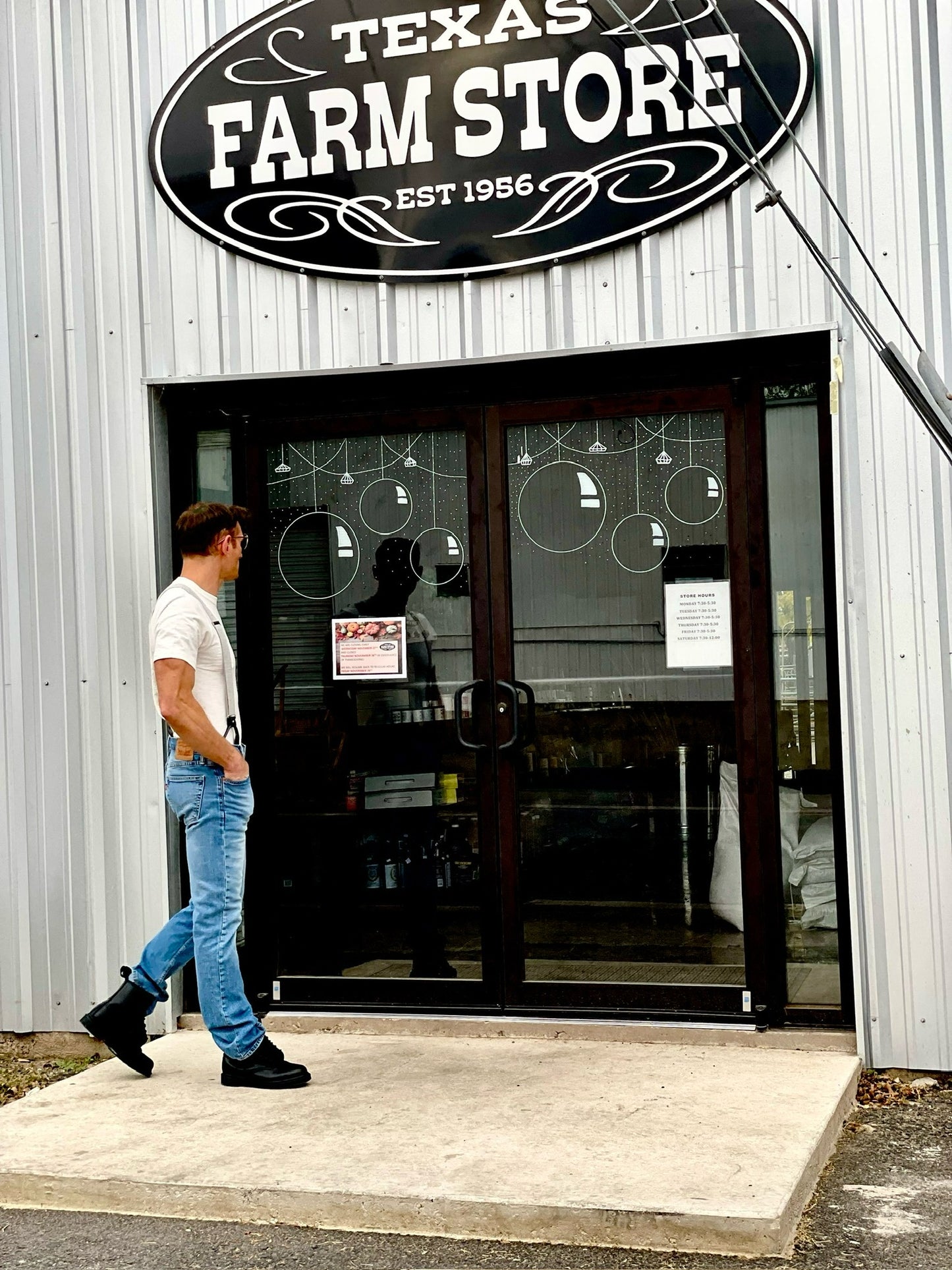 Man in vintage-style suspenders, jeans, and boots walking past Texas Farm Store—Stratton Suspender Co. menswear for rustic weddings or everyday classic style.