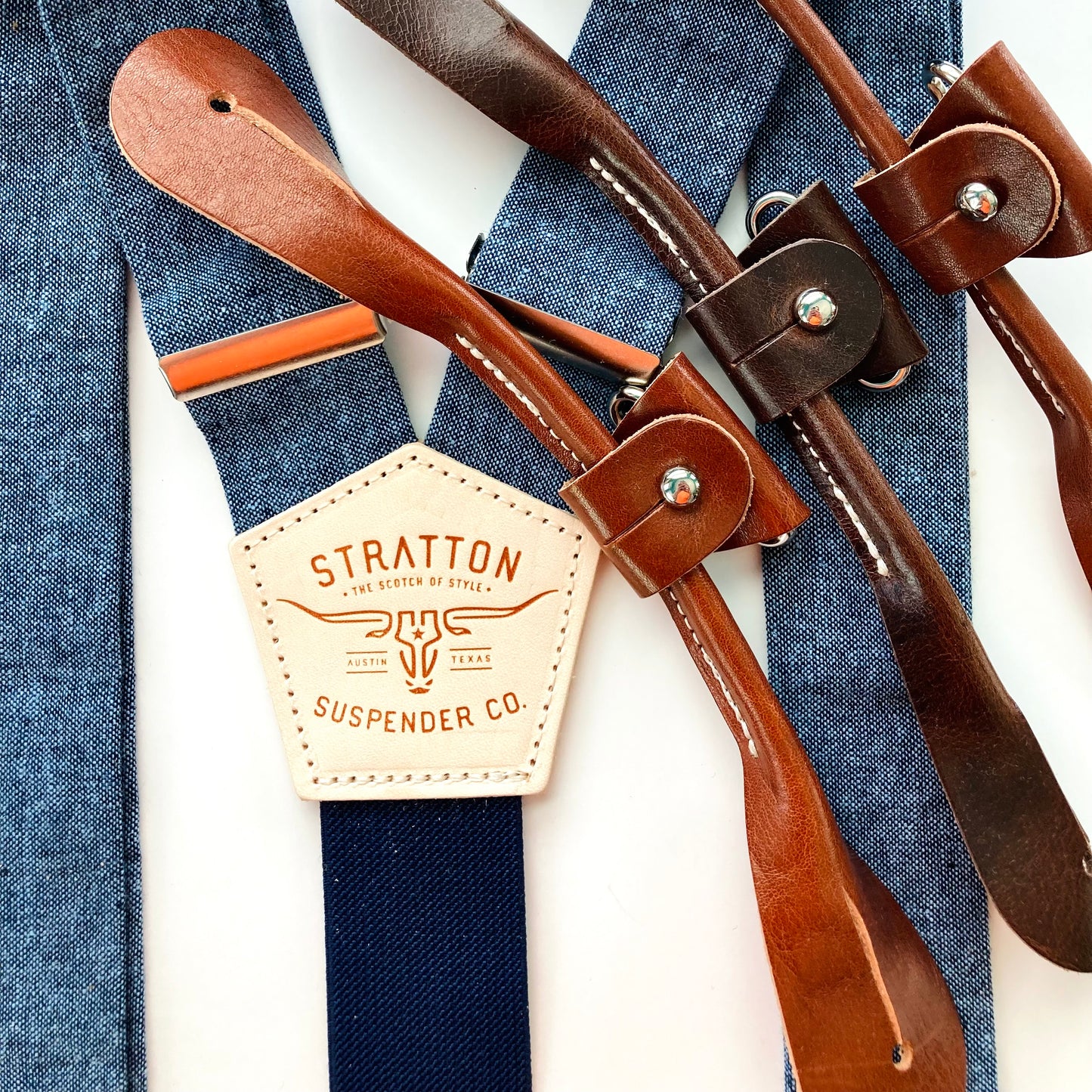 Close-up of Stratton Suspender Co. Nautical Blue linen suspenders featuring the branded leather patch, navy elastic back, and full-grain leather button-on tab options in saddle tan, chestnut, and dark brown.
