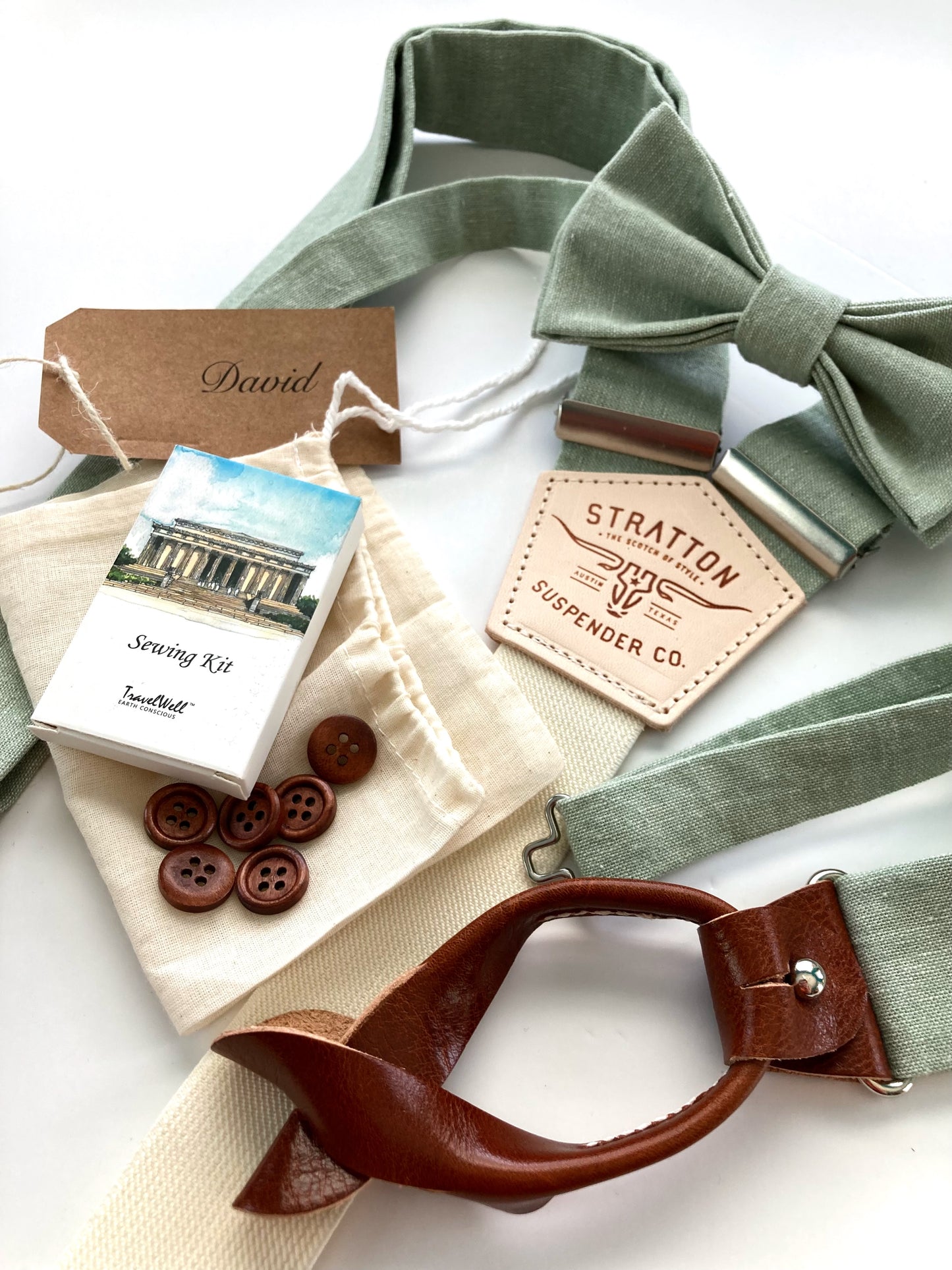 Sage green bow tie and suspenders with leather button tabs, sewing kit, extra buttons, and custom kraft name tag, styled as a groomsmen gift set.