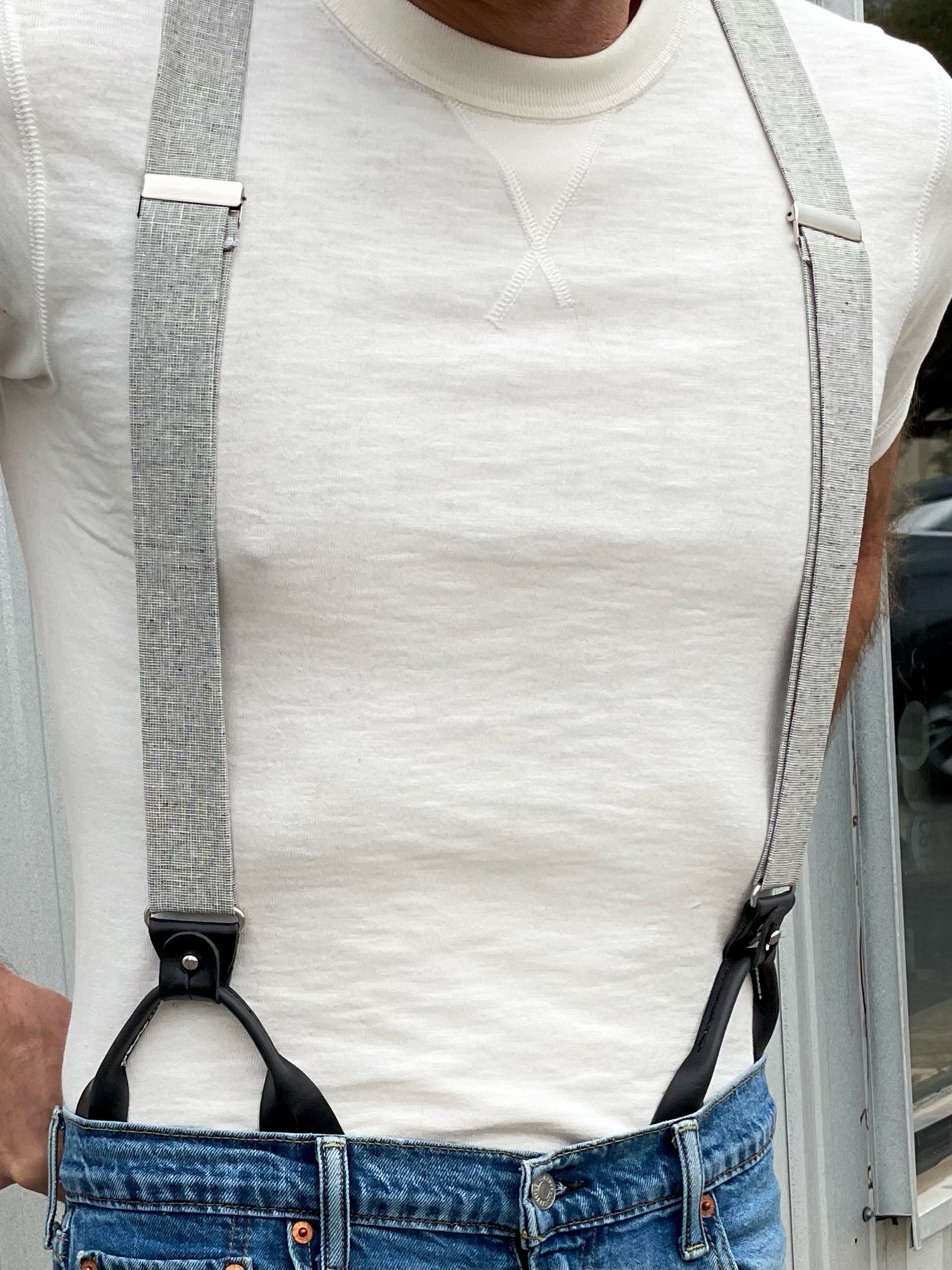Close-up of silver linen suspenders with black full-grain leather button-on attachments, styled with jeans and a cream textured T-shirt. Handmade American men’s suspenders designed for rustic style, modern heritage outfits, and elevated casualwear.