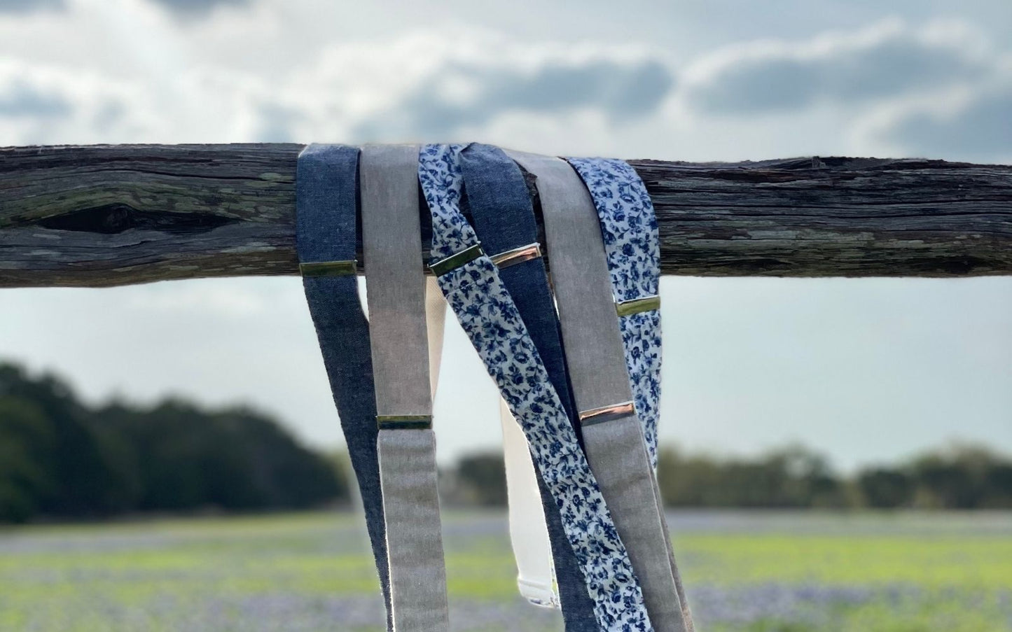 Handmade linen suspenders displayed on a rustic fence overlooking a Texas bluebonnet field and sky.