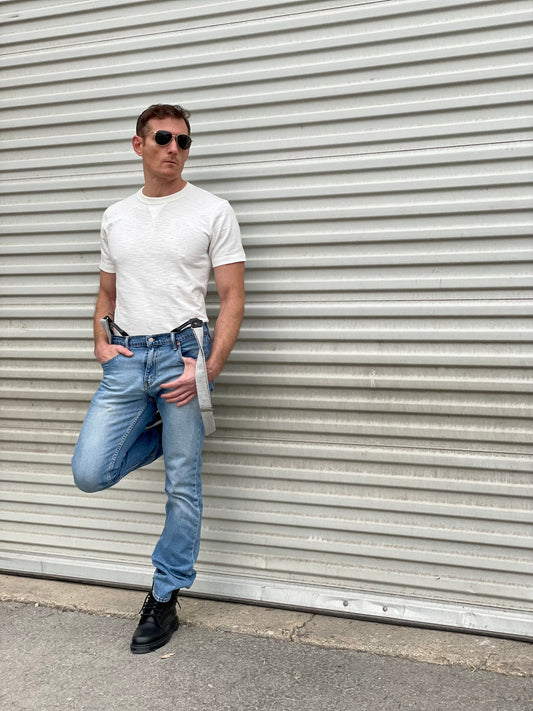 Man wearing a vintage-inspired 1940s heavyweight white t-shirt and wool suspenders by Stratton Suspender Co. Styled with classic denim and black boots, showcasing timeless American workwear against an industrial metal backdrop.