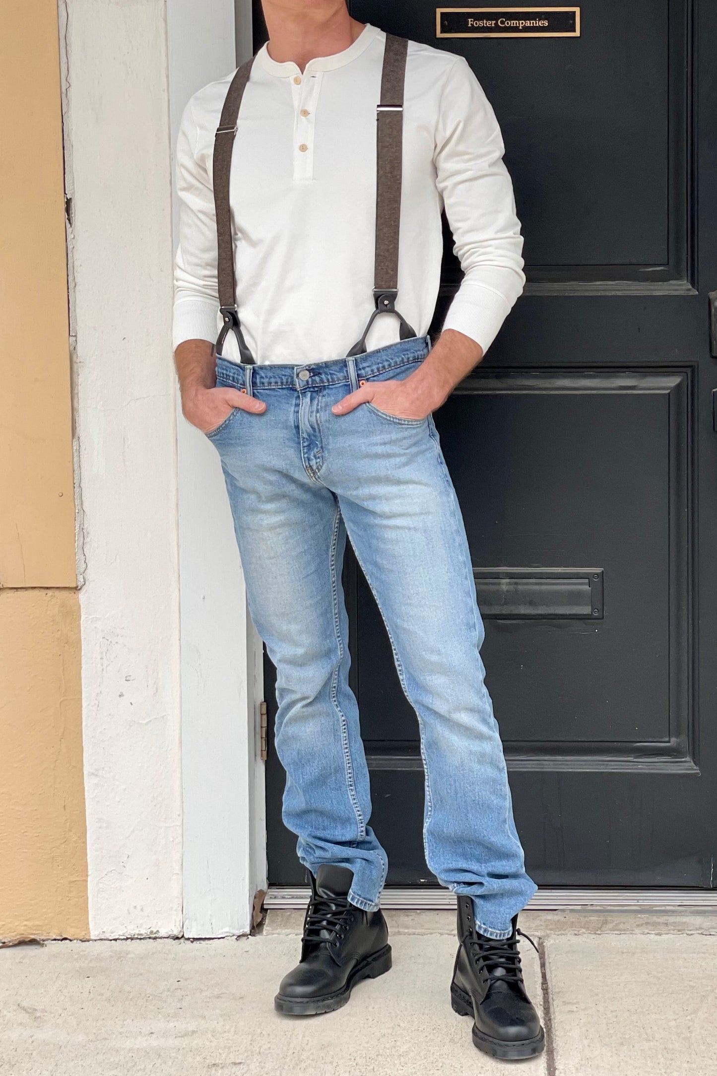 Man wearing a Vintage White Henley and brown wool suspenders by Stratton Suspender Co., standing confidently in front of a black door. Paired with denim and black boots, this rugged heritage menswear look embodies classic American workwear and craftsmanship.