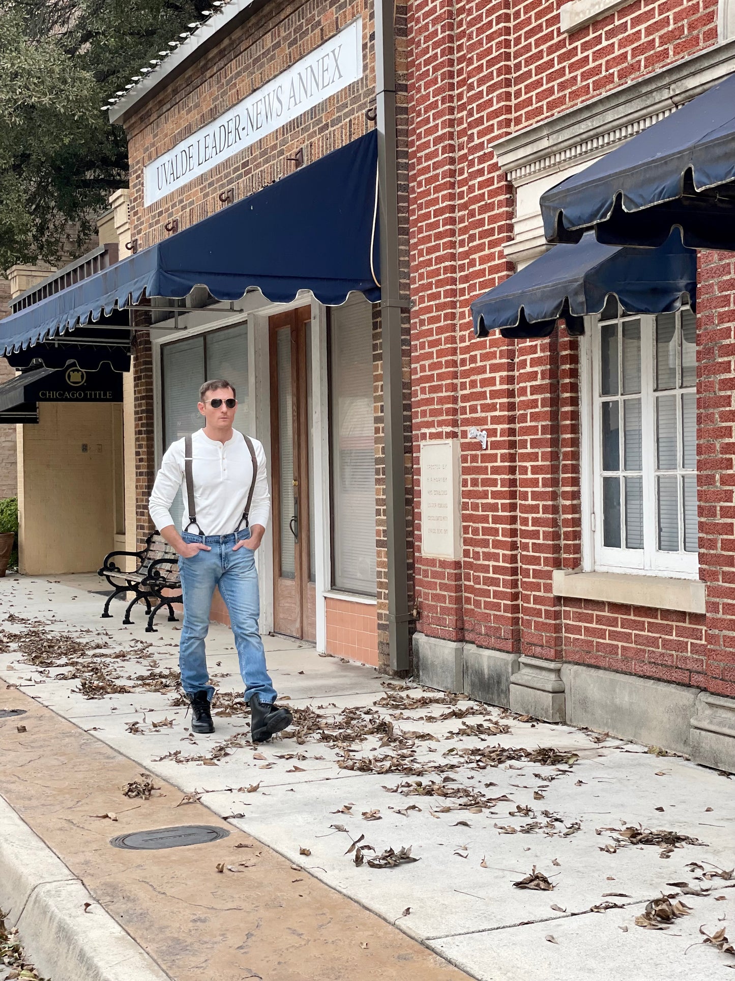 Man walking past a historic brick building wearing a Vintage White Henley and brown wool suspenders by Stratton Suspender Co. Paired with denim and boots, this classic American workwear look captures heritage craftsmanship and timeless menswear style.