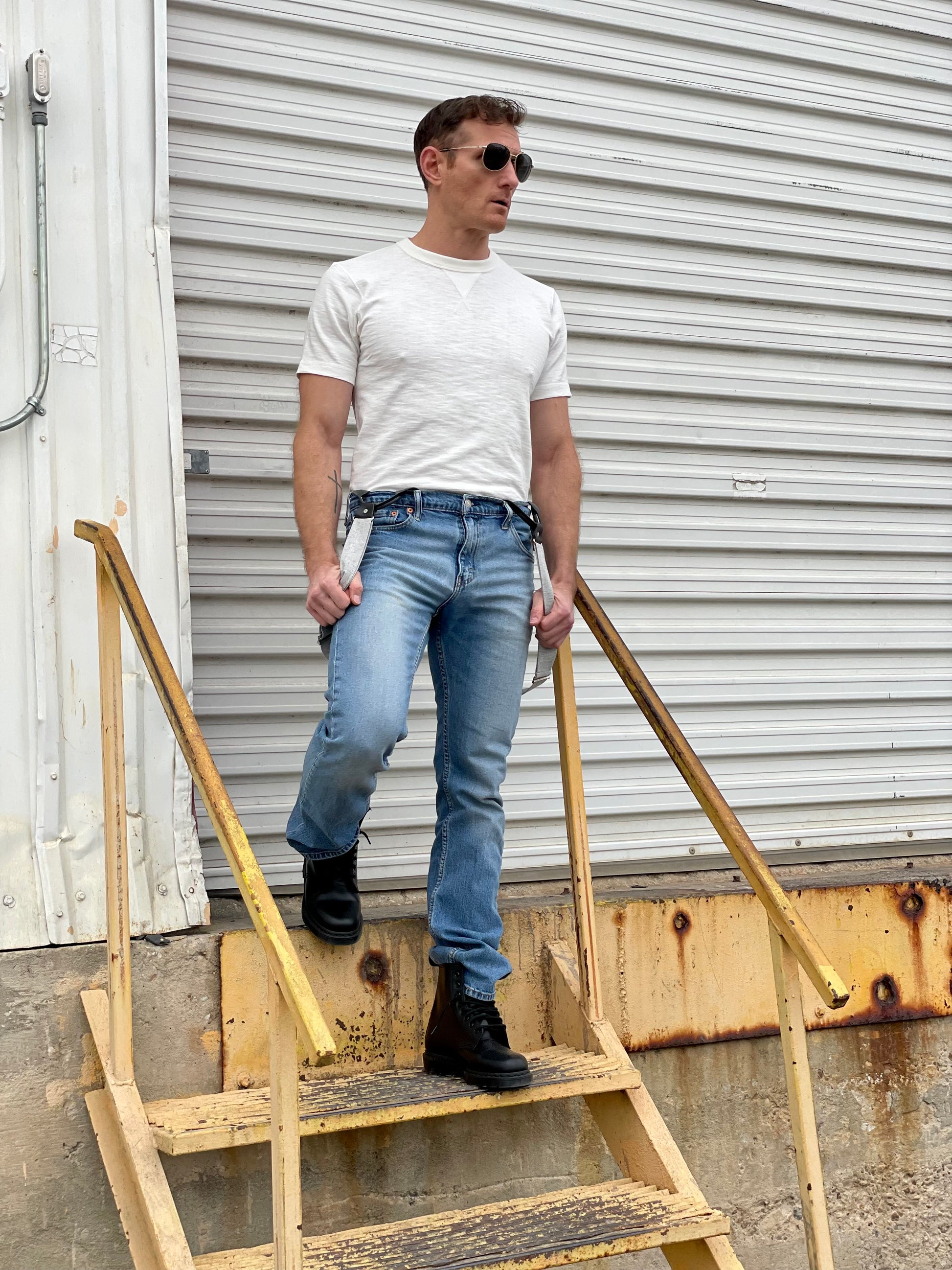 Man wearing a vintage 1940s heavyweight white t-shirt with gray linen suspenders by Stratton Suspender Co. Styled with classic denim and rugged black boots, he steps down a weathered industrial staircase, capturing timeless American workwear and heritage menswear in an authentic setting.