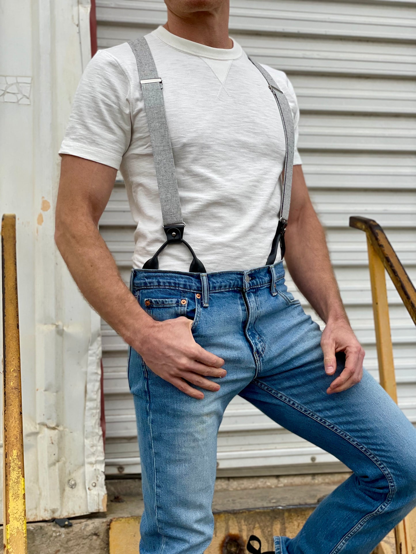 Close-up of a man wearing a vintage 1940s heavyweight white t-shirt and gray linen suspenders by Stratton Suspender Co. Paired with classic blue jeans, the rugged industrial setting highlights the timeless appeal of heritage American workwear and handcrafted menswear.