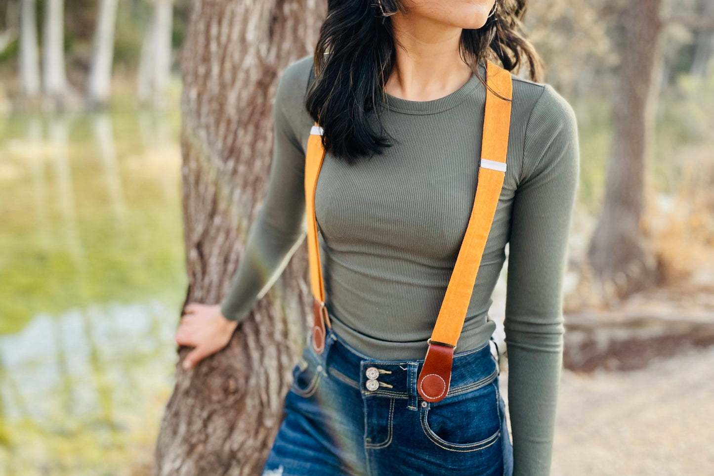 Close-up of women’s tangerine orange linen suspenders with tan leather magnetic clasps styled with dark denim jeans and fitted ribbed top, photographed outdoors by a tree near water.