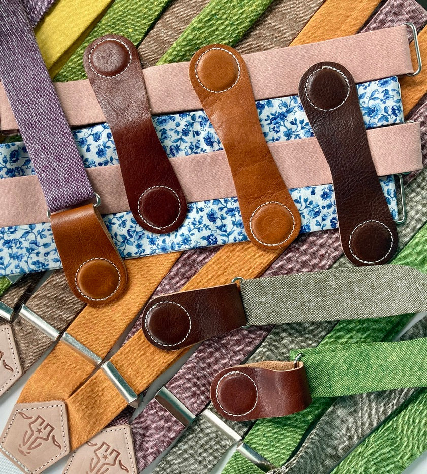 Flat-lay of women’s linen suspenders in bold fall colors—purple, blush, olive, kelly green, rust, nutmeg, orange, and blue floral—with cognac, tan, and chocolate leather magnetic clasps.
