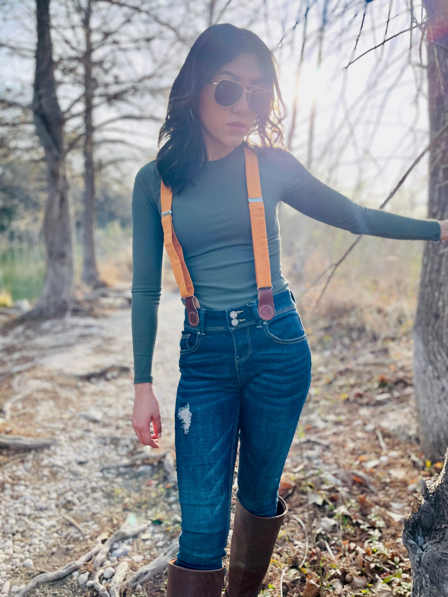 Women’s tangerine orange linen suspenders with tan magnetic clasps styled with high-rise jeans, boots, and fitted top in a rustic fall outdoor setting.