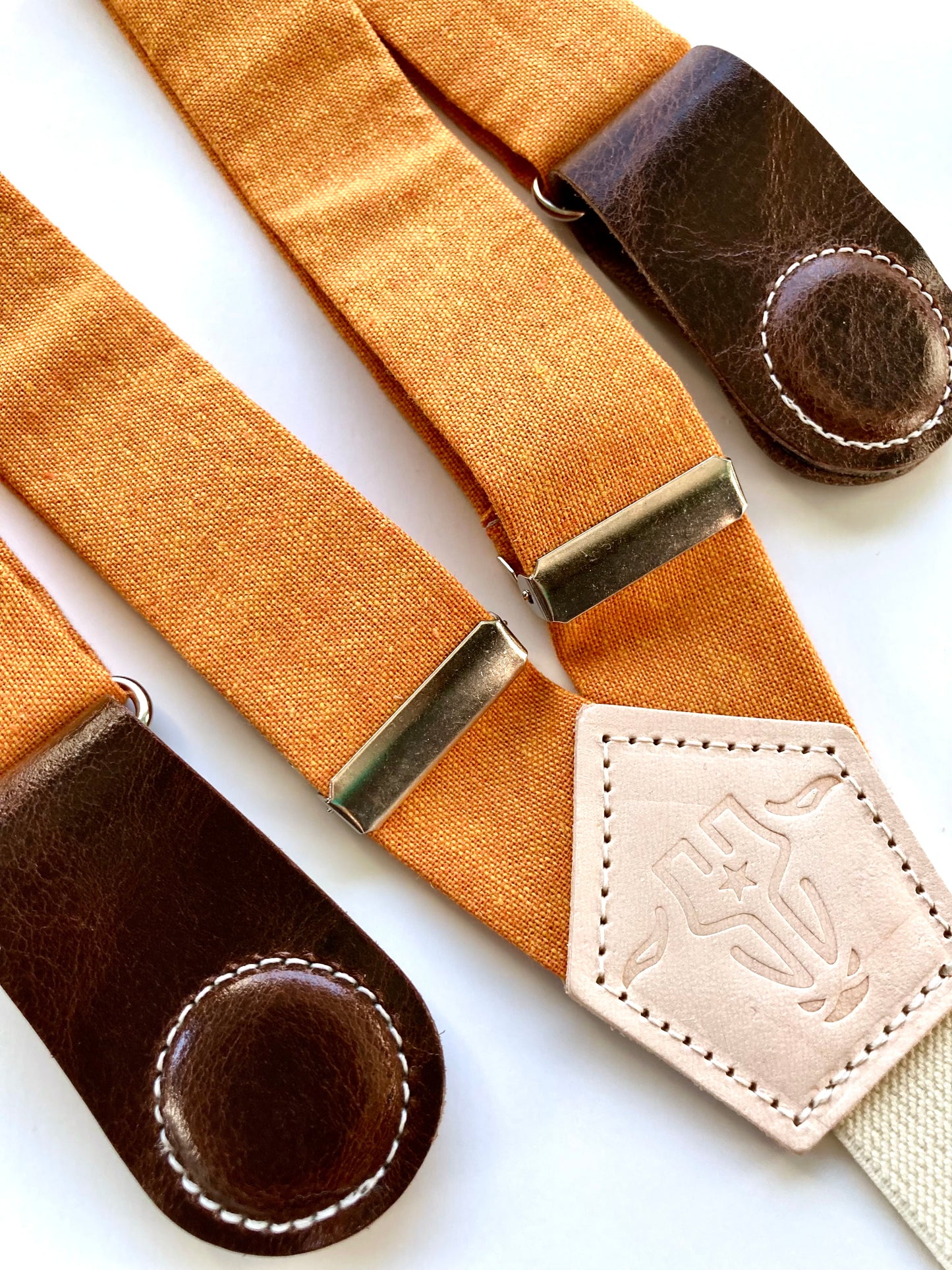 Detail of women’s tangerine orange linen suspenders with chocolate leather magnetic clasps on white background.