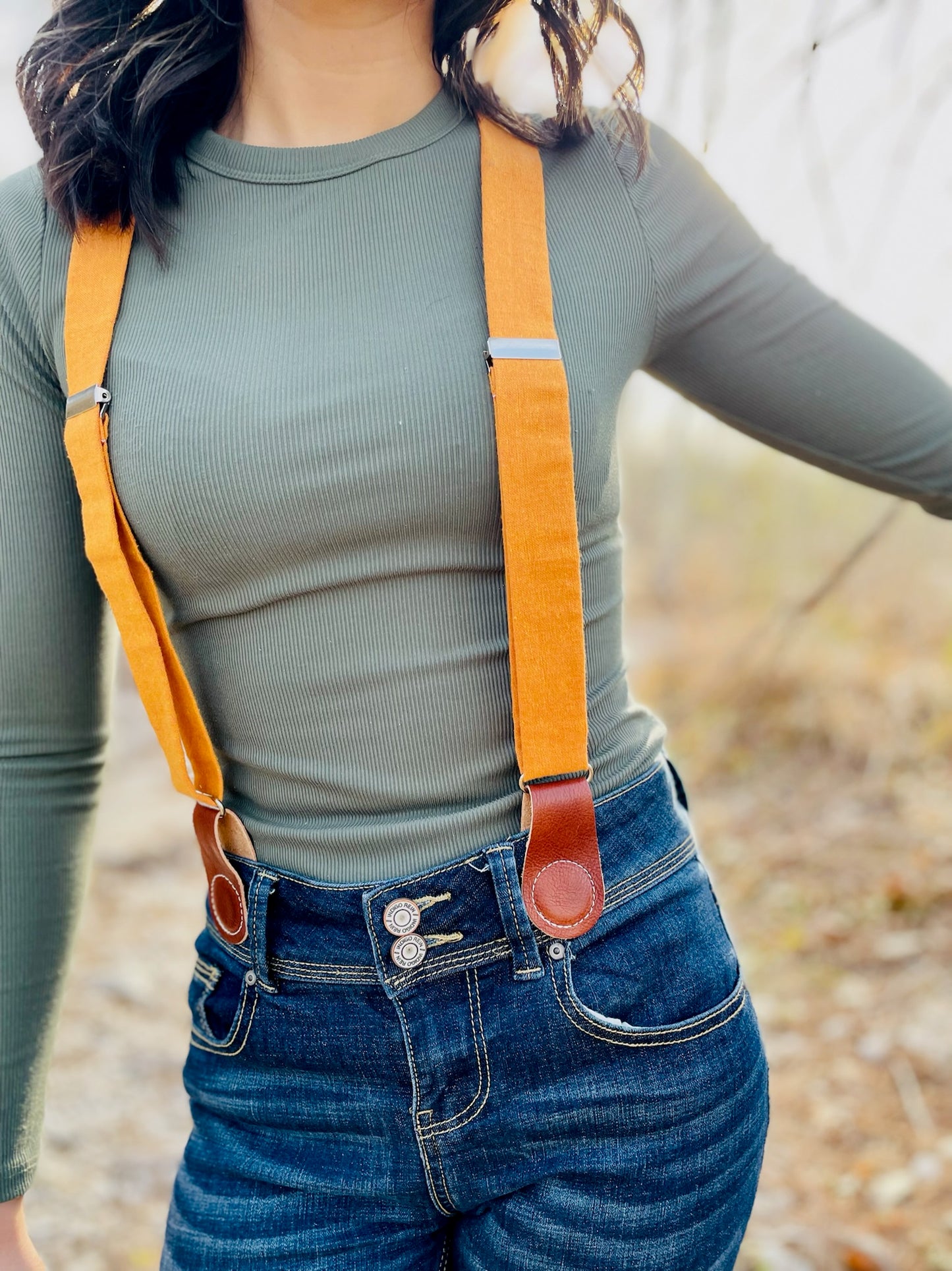 Close-up of women’s tangerine orange linen suspenders with tan leather magnetic clasps styled with high-waist jeans and ribbed top.