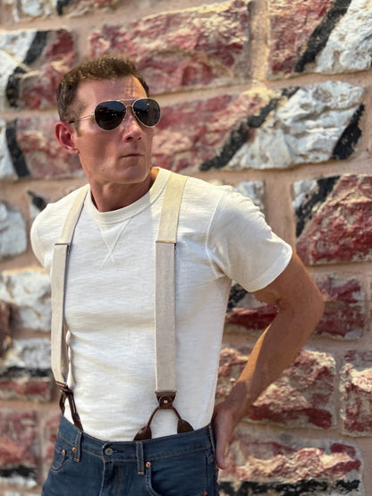 A man wearing a white crewneck t-shirt, beige canvas suspenders with brown leather loops, and blue jeans, standing in front of a rustic red and gray stone wall.