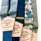 A collection of blue linen and floral suspenders with navy, cream, and gray elastic back straps. Features premium leather back patches and gold-tone adjusters. Handmade suspenders for weddings, groomsmen, and vintage menswear. Ethically crafted for a timeless, classic look.