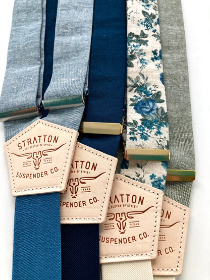A collection of blue linen and floral suspenders with navy, cream, and gray elastic back straps. Features premium leather back patches and gold-tone adjusters. Handmade suspenders for weddings, groomsmen, and vintage menswear. Ethically crafted for a timeless, classic look.