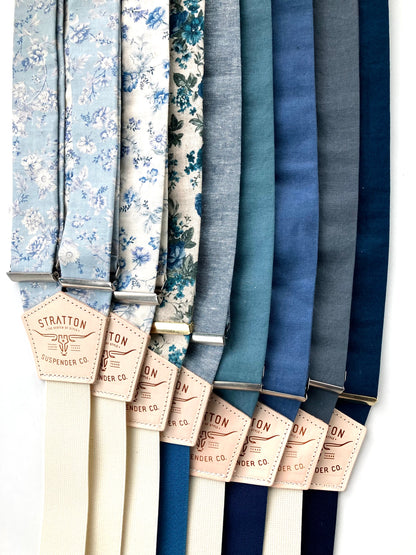 A collection of blue linen and floral wedding suspenders with cream and navy elastic back straps. Features premium vegetable-tanned leather back patches. Handmade wedding suspenders, perfect for groomsmen, rustic weddings, and heritage menswear. Ethically crafted for timeless style.