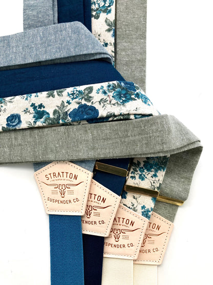 A collection of blue linen and floral wedding suspenders with deep blue, cream, and gray elastic back straps. Features handcrafted leather back patches and gold-tone adjusters. Premium suspenders for rustic weddings, groomsmen, and heritage menswear. Ethically made for a classic, timeless style.