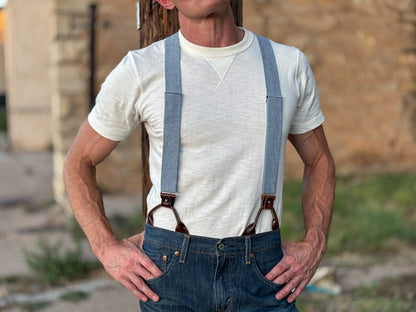A man wearing light blue denim suspenders with brown leather hardware, styled over a white t-shirt and blue jeans.