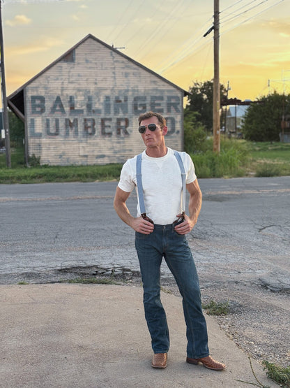 A man wearing a white t-shirt, blue denim jeans, light blue suspenders, and brown leather shoes stands on a paved road in front of a rustic building with 'BALLINGER LUMBER CO' painted on it.