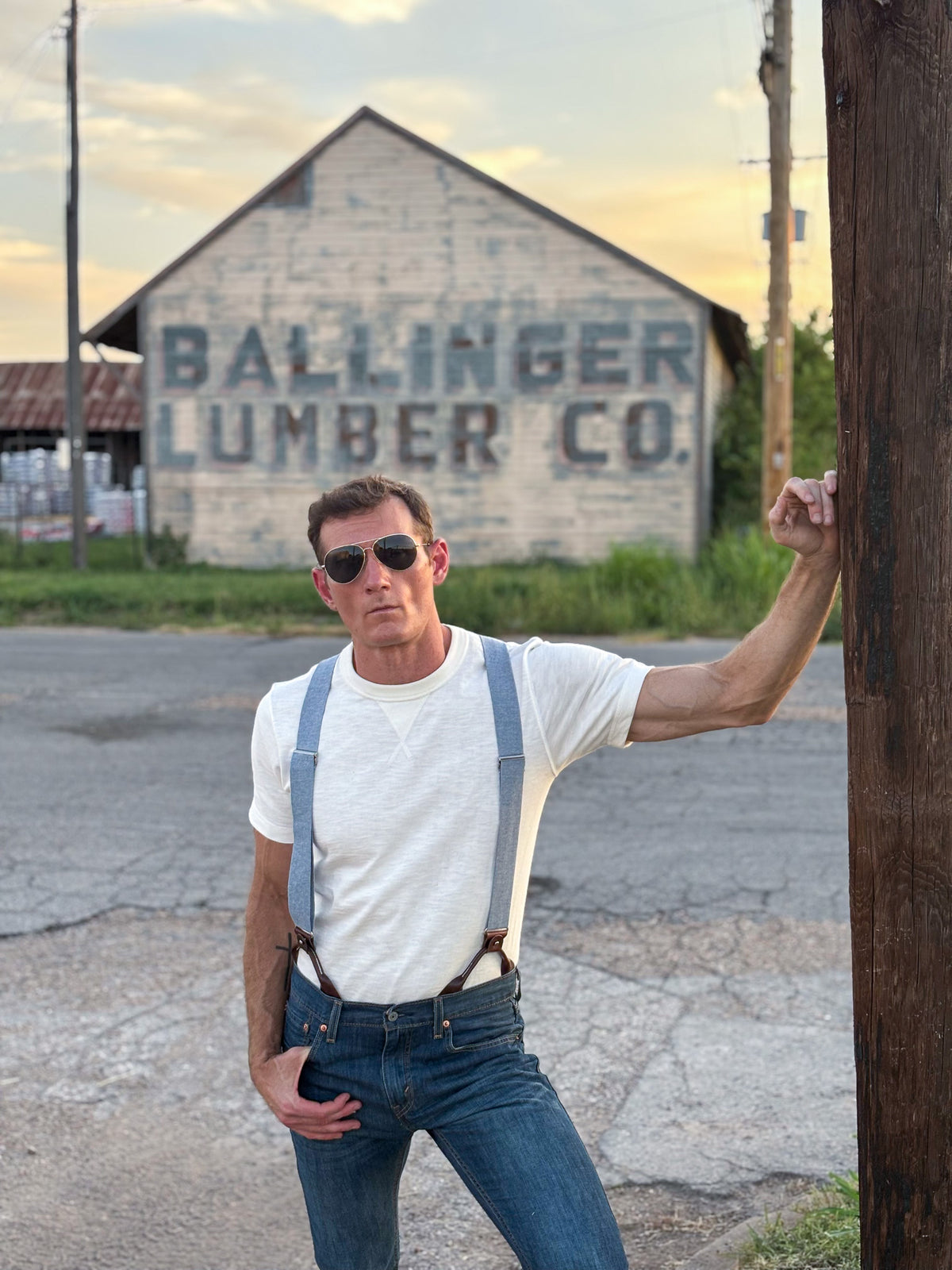 A man wearing a white t-shirt, blue jeans, and blue suspenders stands outdoors in front of a rustic building with 'BALLINGER LUMBER CO.' painted on the side, holding onto a wooden utility pole.