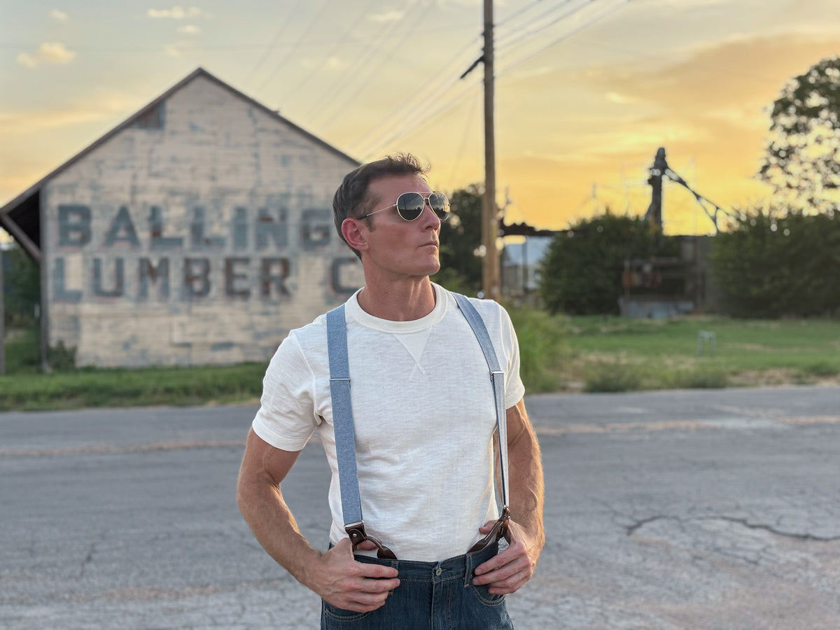 A man wearing a white t-shirt, light blue denim suspenders, and dark jeans stands on an asphalt road in front of a rustic building with 'BALLING LUMBER CO' painted on it, during sunset.