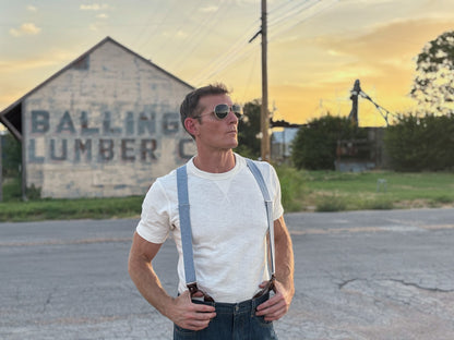 A man wearing a white t-shirt, light blue denim suspenders, and dark jeans stands on an asphalt road in front of a rustic building with 'BALLING LUMBER CO' painted on it, during sunset.
