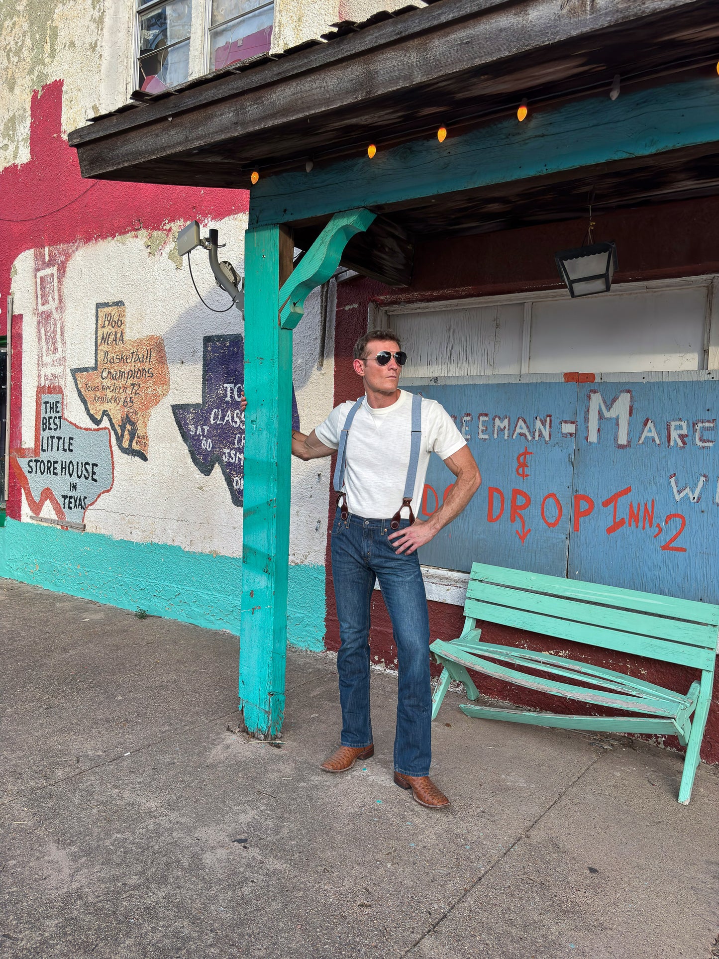 A man wearing a white t-shirt, blue jeans, and blue suspenders stands under a teal wooden shelter on a sidewalk in front of a colorful graffiti wall.