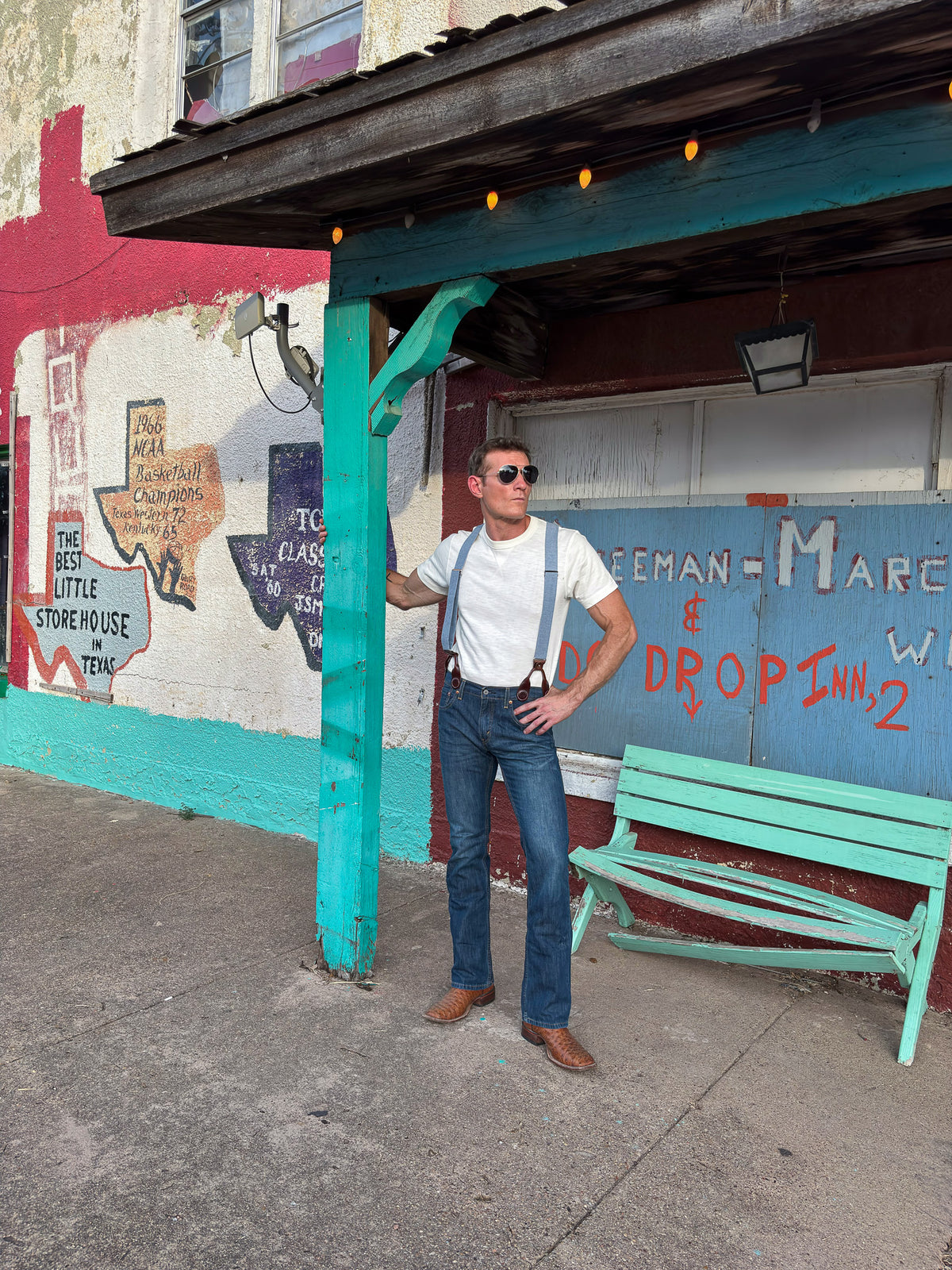 A man wearing a white t-shirt, blue jeans, and blue suspenders stands under a teal wooden shelter on a sidewalk in front of a colorful graffiti wall.
