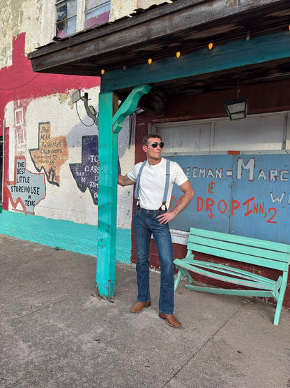 A man wearing a white t-shirt, blue jeans, and blue suspenders stands under a teal wooden shelter on a sidewalk in front of a colorful graffiti wall.