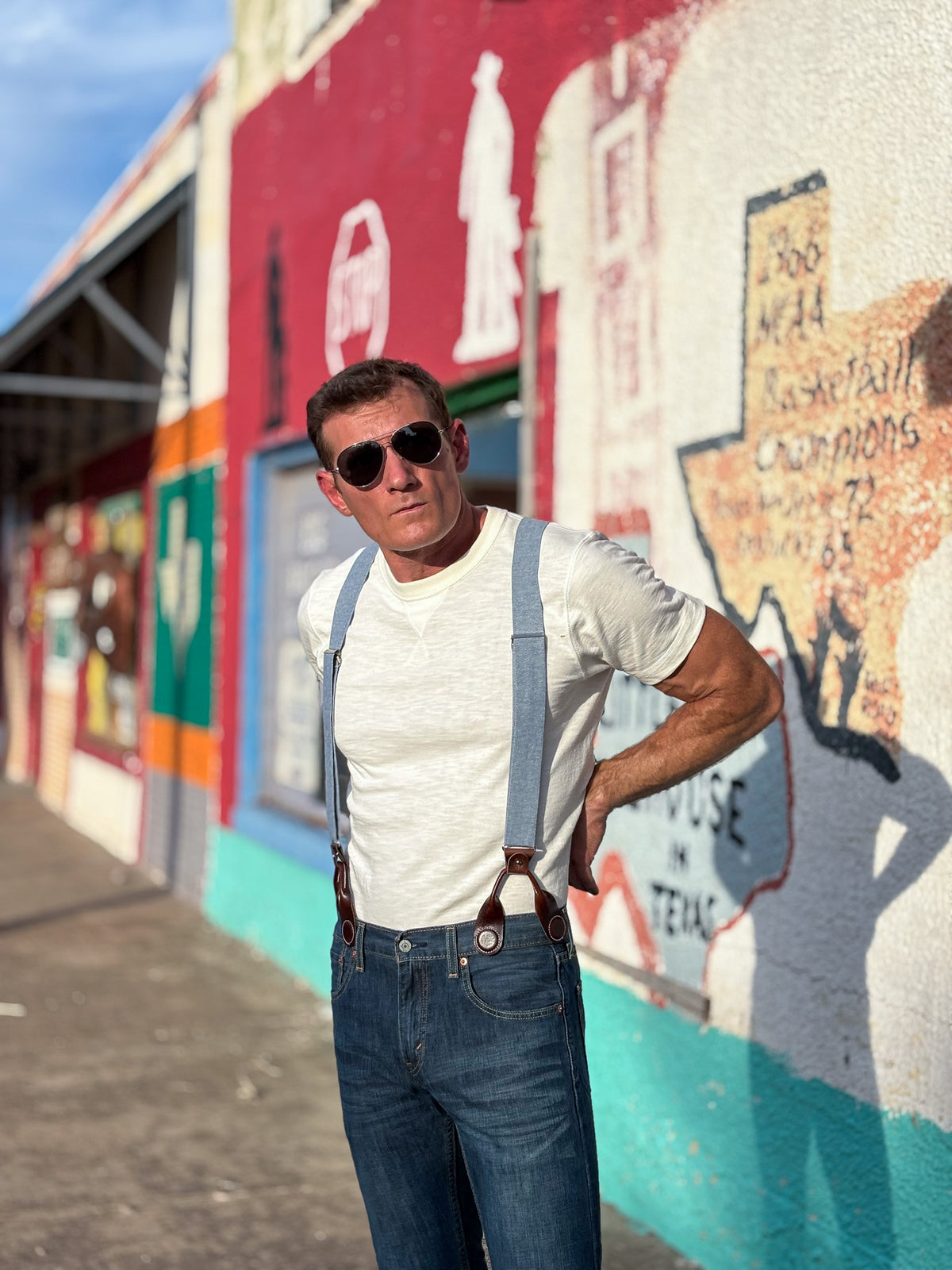 A man wearing a white t-shirt, blue jeans, and light blue suspenders stands in front of a colorful painted wall with murals.
