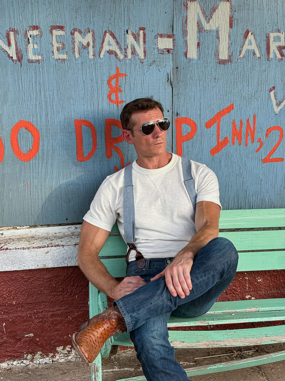 A man wearing aviator sunglasses, a white t-shirt, blue jeans, and gray suspenders sits on a distressed green wooden bench in front of a weathered blue painted wall with orange and white painted text.